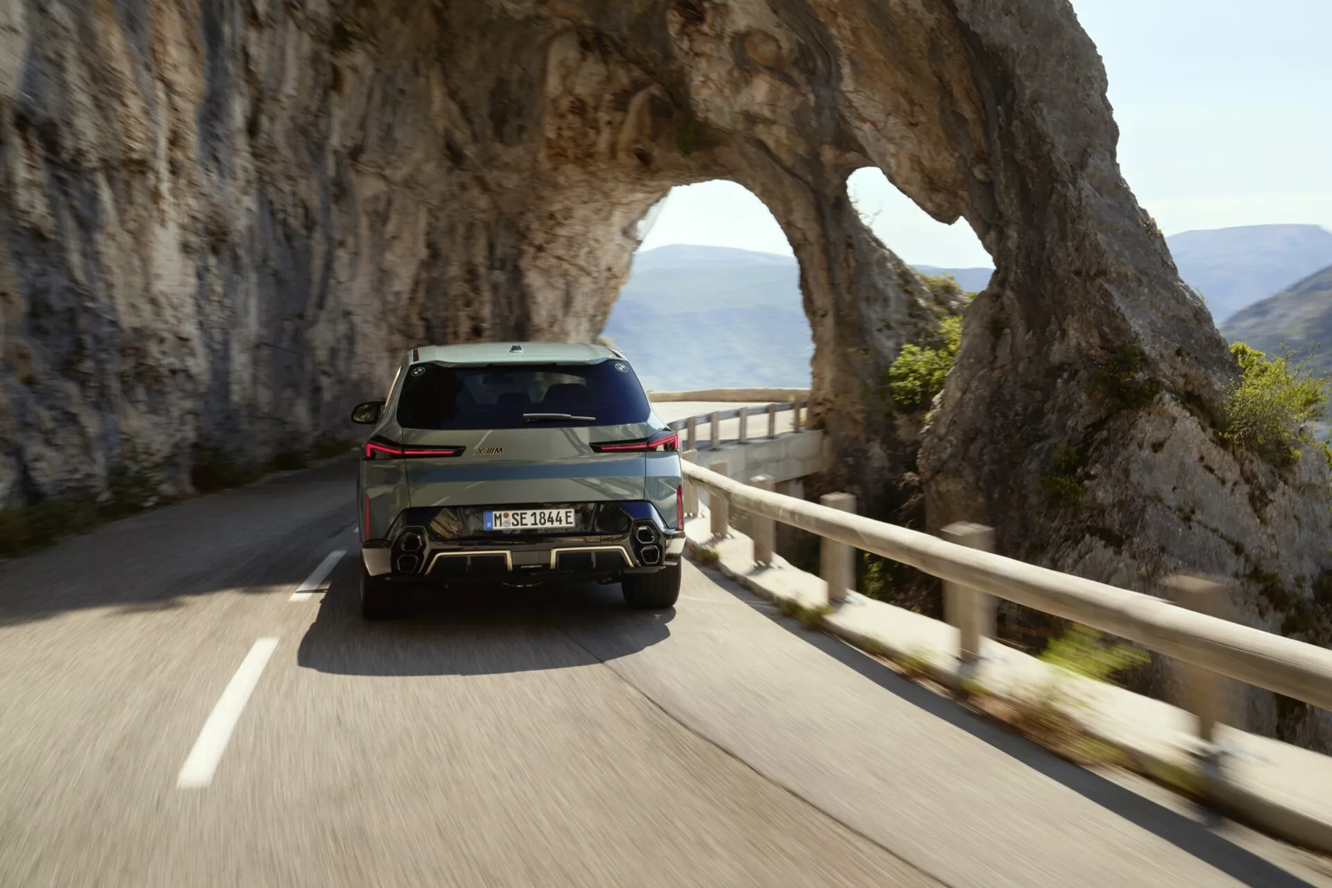 2023 BMW XM (G09) driving on a mountainous road with natural arch, rear three-quarter view highlighting LED taillights, bumper design, and a glimpse of its sleek side profile in morning soft light