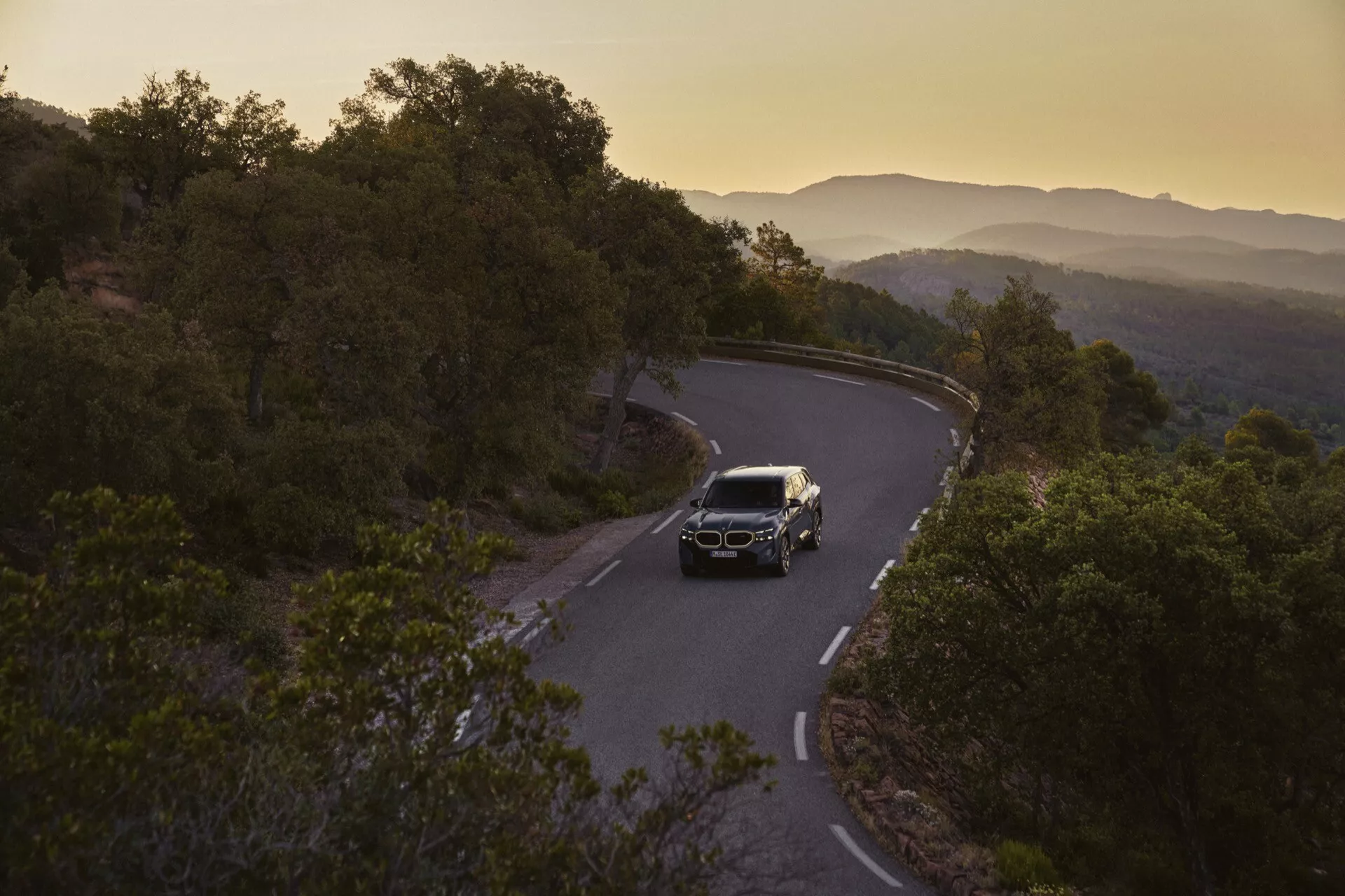 2023 BMW XM (G09) luxury SUV driving on a winding road at dawn or dusk, showcasing its LED headlights, prominent grille, and alloy wheels amidst a scenic landscape