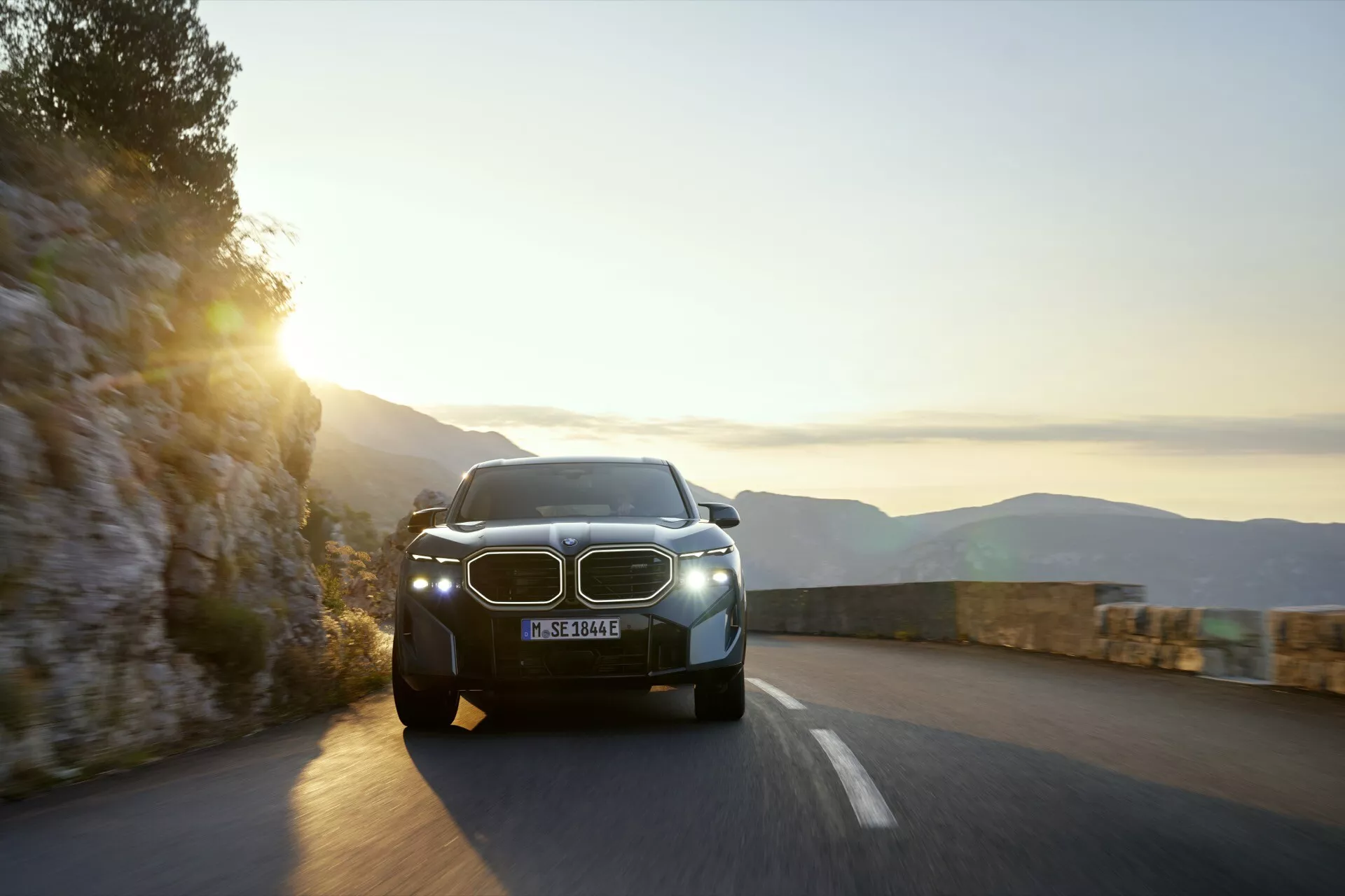 2023 BMW XM (G09) modern SUV driving on a mountain road at dawn or dusk with illuminated LED headlights and distinctive kidney-shaped grille visible