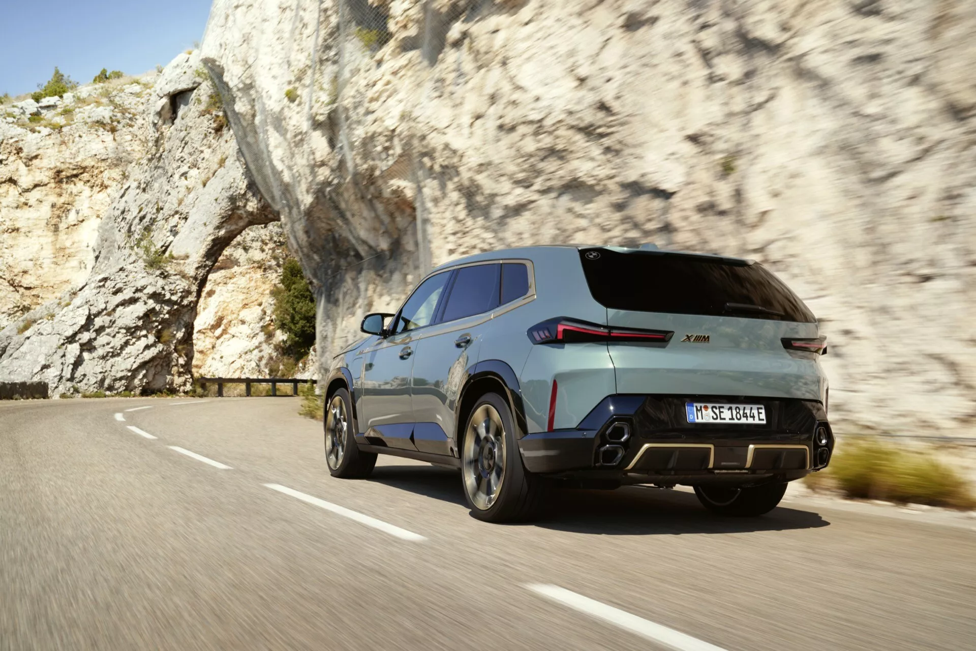 Dynamic rear three-quarter view of a modern SUV, possibly the 2023 BMW XM (G09), driving on a mountain road showcasing the right rear wheel, taillight, tailgate, and portion of the right side body panel towards the front door, with visible motion blur, dual exhaust system, roof spoiler, and large alloy wheels with a metallic finish, surrounded by natural rocky formations.