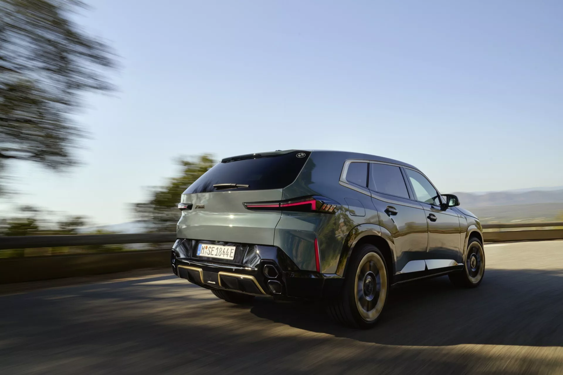Dynamic low angle shot of the 2023 BMW XM (G09) in motion, featuring rear three-quarter view with LED tail lights, dual exhaust, and roof spoiler, showcasing speed and modern design.