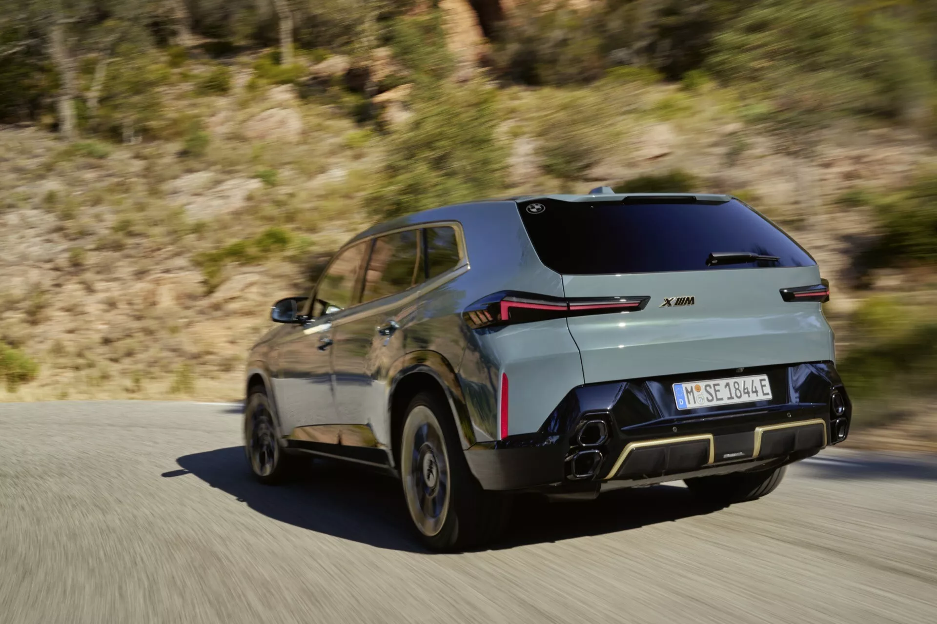 2023 BMW XM (G09) modern SUV in motion on a paved road, featuring sporty aerodynamic design, LED taillights, dual-tone body, reflective parking sensors, polygonal dual exhaust, alloy wheels, and potential electric/hybrid badge, set against a rural/mountainous backdrop