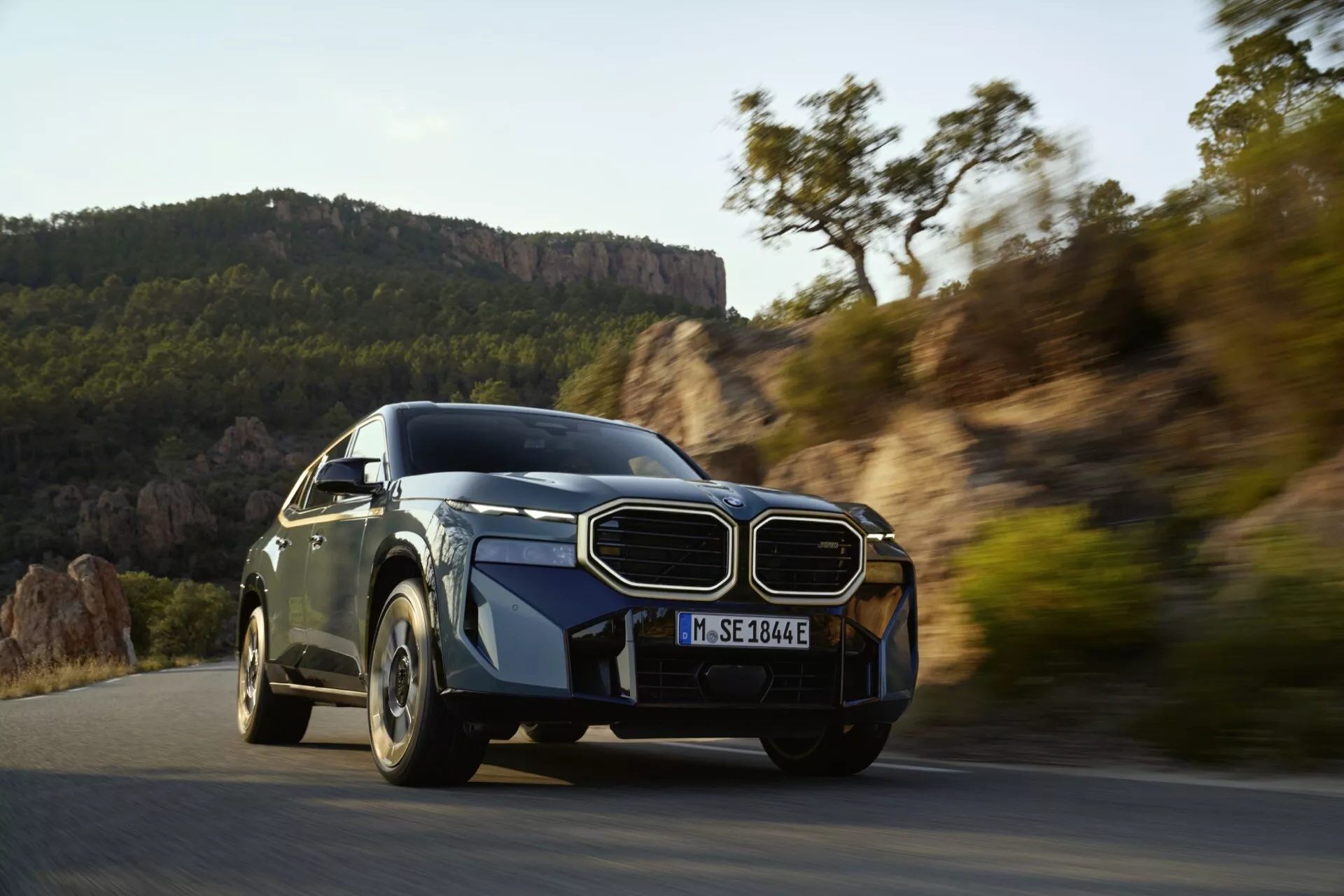 Dynamic capture of a luxury vehicle in motion, showcasing its advanced aerodynamic design, prominent grille, sleek LED headlights, and sporty multi-spoke wheels against a scenic outdoor backdrop