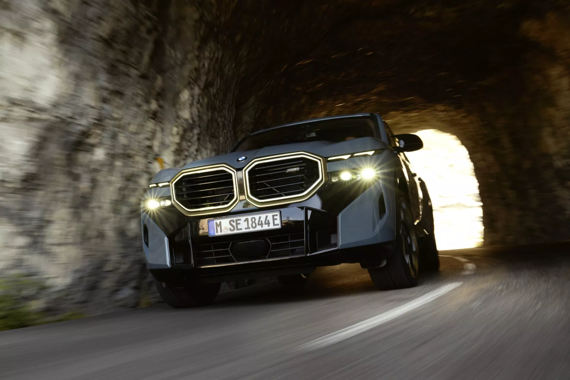 2023 BMW XM (G09) exiting tunnel, low-angle view showcasing grille, LED headlights, and hood design contrasted by daylight and tunnel shadow