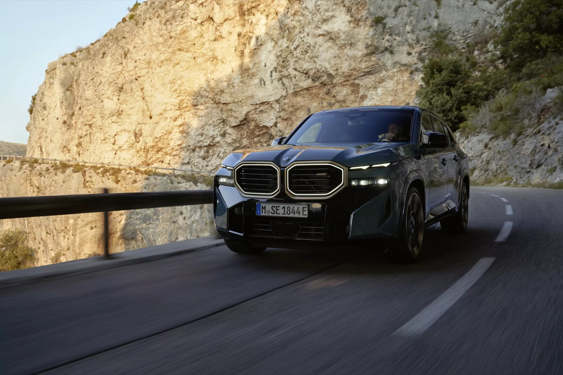 2023 BMW XM (G09) in motion on a winding mountain road, showcasing its dynamic design and advanced technological features like the iconic kidney-shaped grille, LED headlights, and alloy wheels