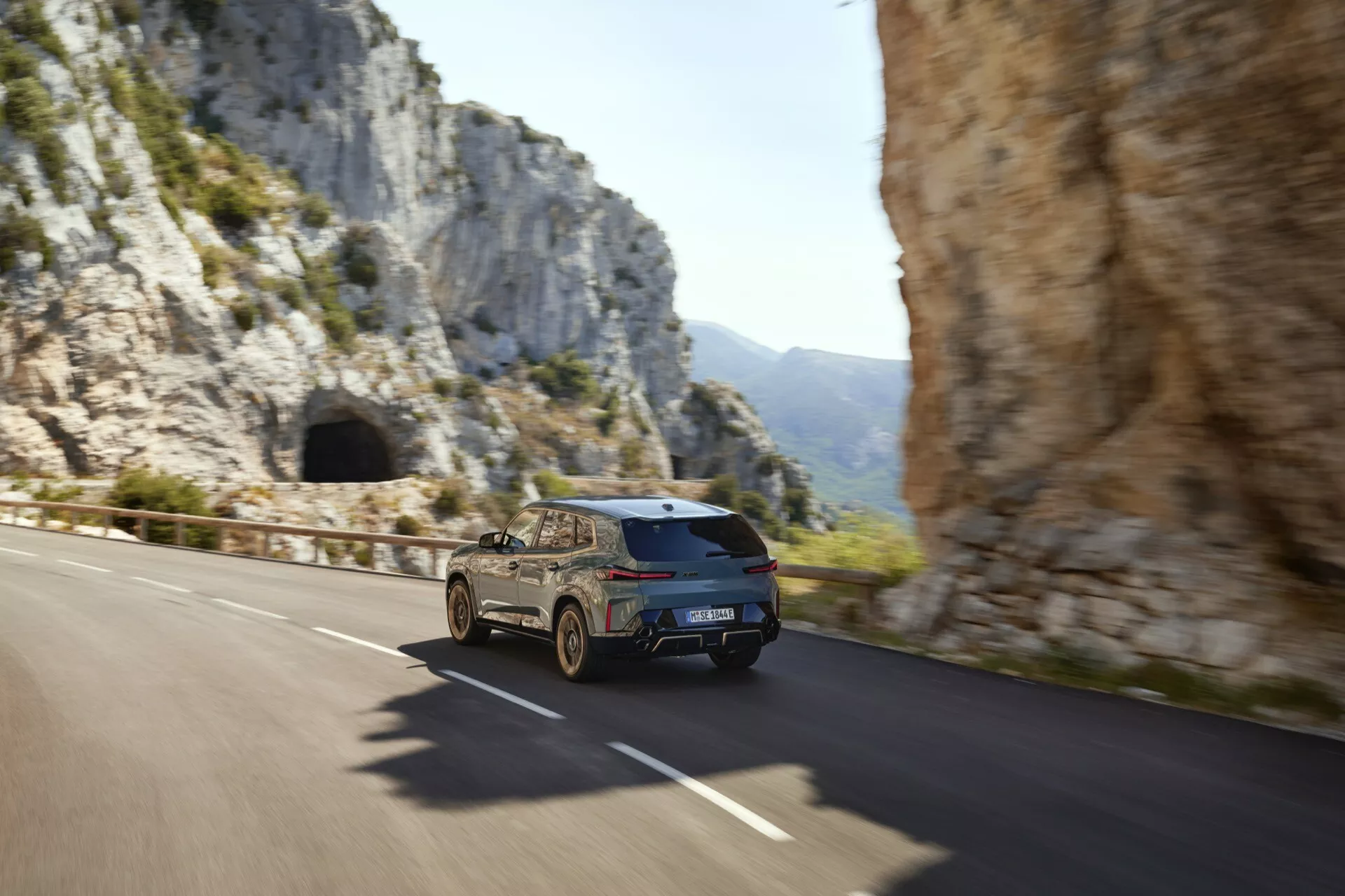 2023 BMW XM (G09) in motion, captured from a rear three-quarter angle showcasing the right rear quarter panel, taillights, trunk, and right rear wheel, against a mountainous backdrop, embodying dynamic movement and advanced LED technology.