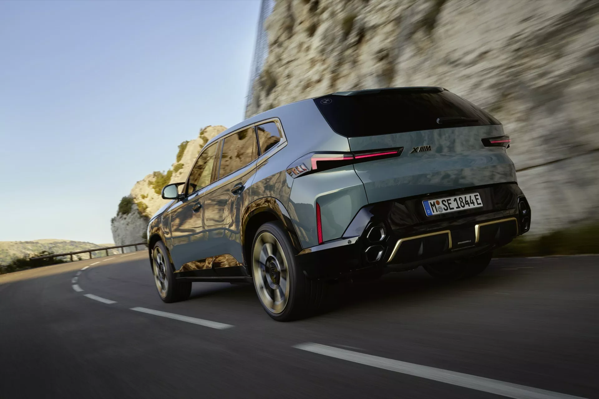 2023 BMW XM (G09) dynamic rear three-quarter view in motion, showcasing its modern SUV design with LED tail lights, AWD badge, dual-tone bumper with integrated parking sensors, silver skid plate accents, multi-spoke alloy wheels, and metallic paint finish in a natural, brightly lit outdoor setting