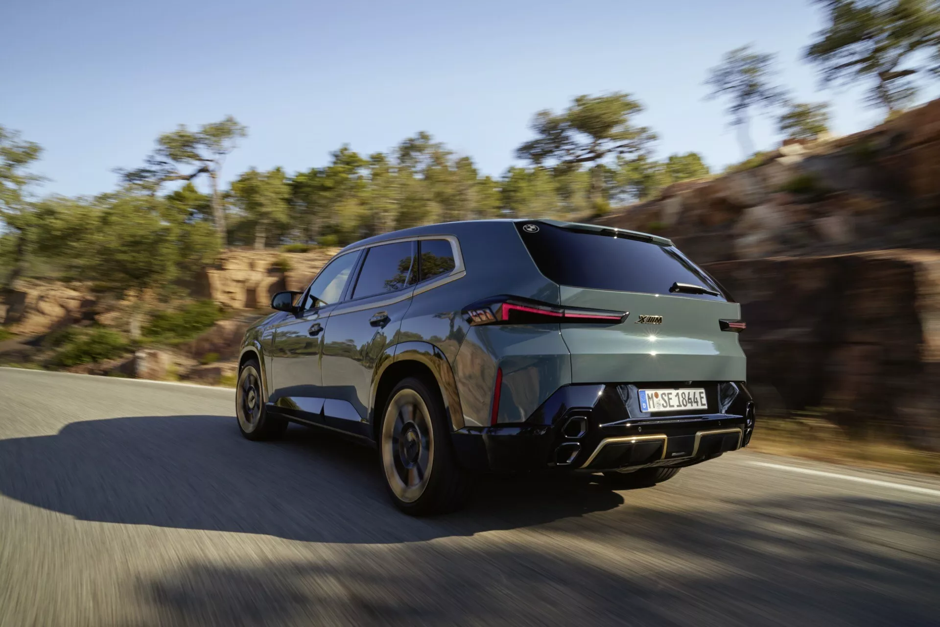 2023 BMW XM (G09) captured in dynamic motion from a rear three-quarter perspective, highlighting its sleek design with LED tail lights, metallic finish, and high-performance wheels on a sunny road.