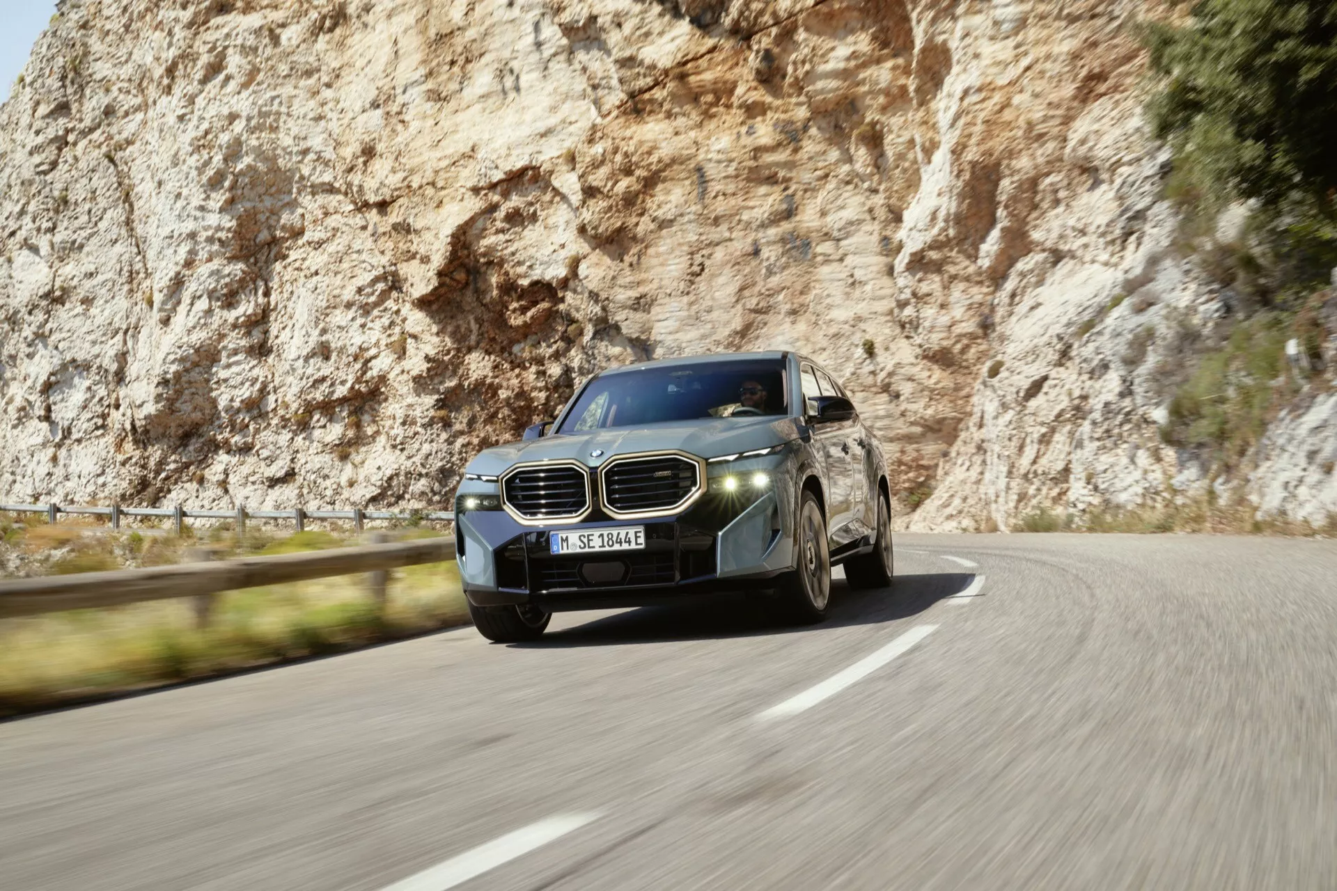 2023 BMW XM (G09) in dynamic motion on a curvy road, showcasing its luxurious front three-quarter profile, distinctive dual kidney-shaped grille, sleek LED headlights, multi-spoke alloy wheels, and aerodynamic body with metallic paint and chrome accents.