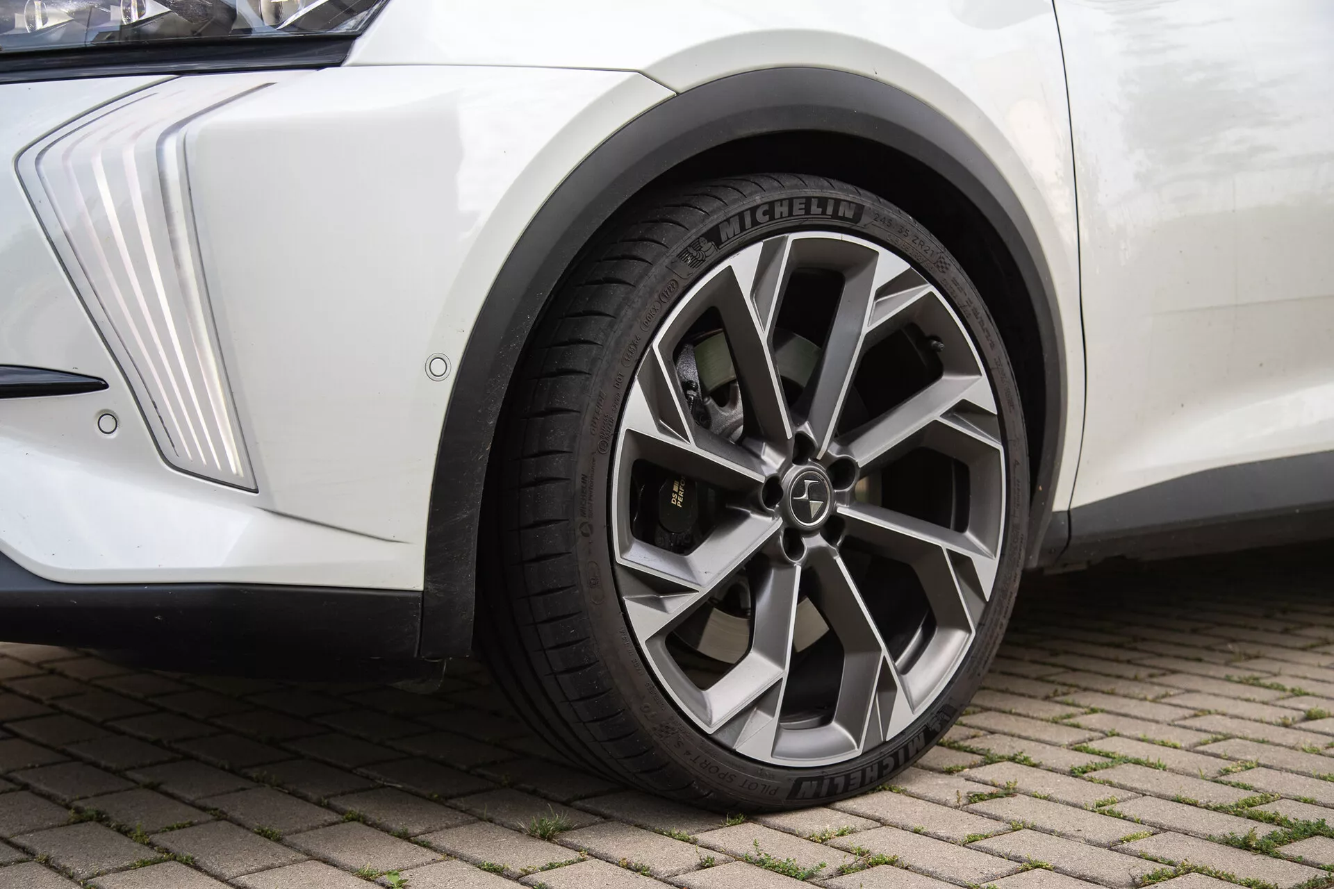Close-up view of a DS-Automobiles DS-7 2022 front wheel assembly on interlocking brick surface, featuring a metallic finish multi-spoke alloy wheel with a central logo cap, Michelin radial tire with detailed tread patterns, angular-designed white fender, and a black mud flap, under natural lighting