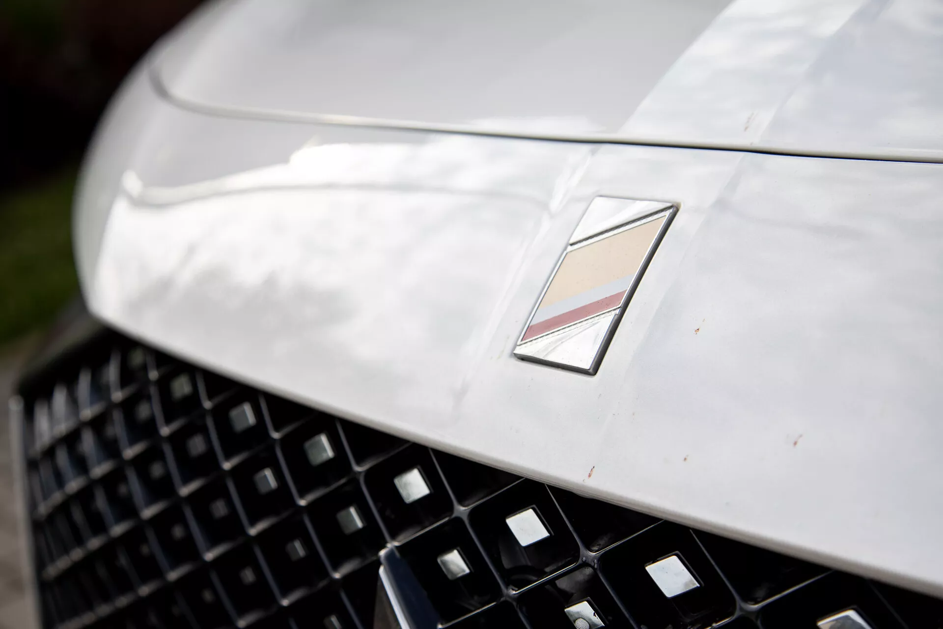 Close-up view of a luxury car's front section showcasing a metallic hood with aerodynamic design, a detailed grille for enhanced air intake, and a multifaceted emblem signifying the manufacturer, under natural light reflection and shallow depth of field