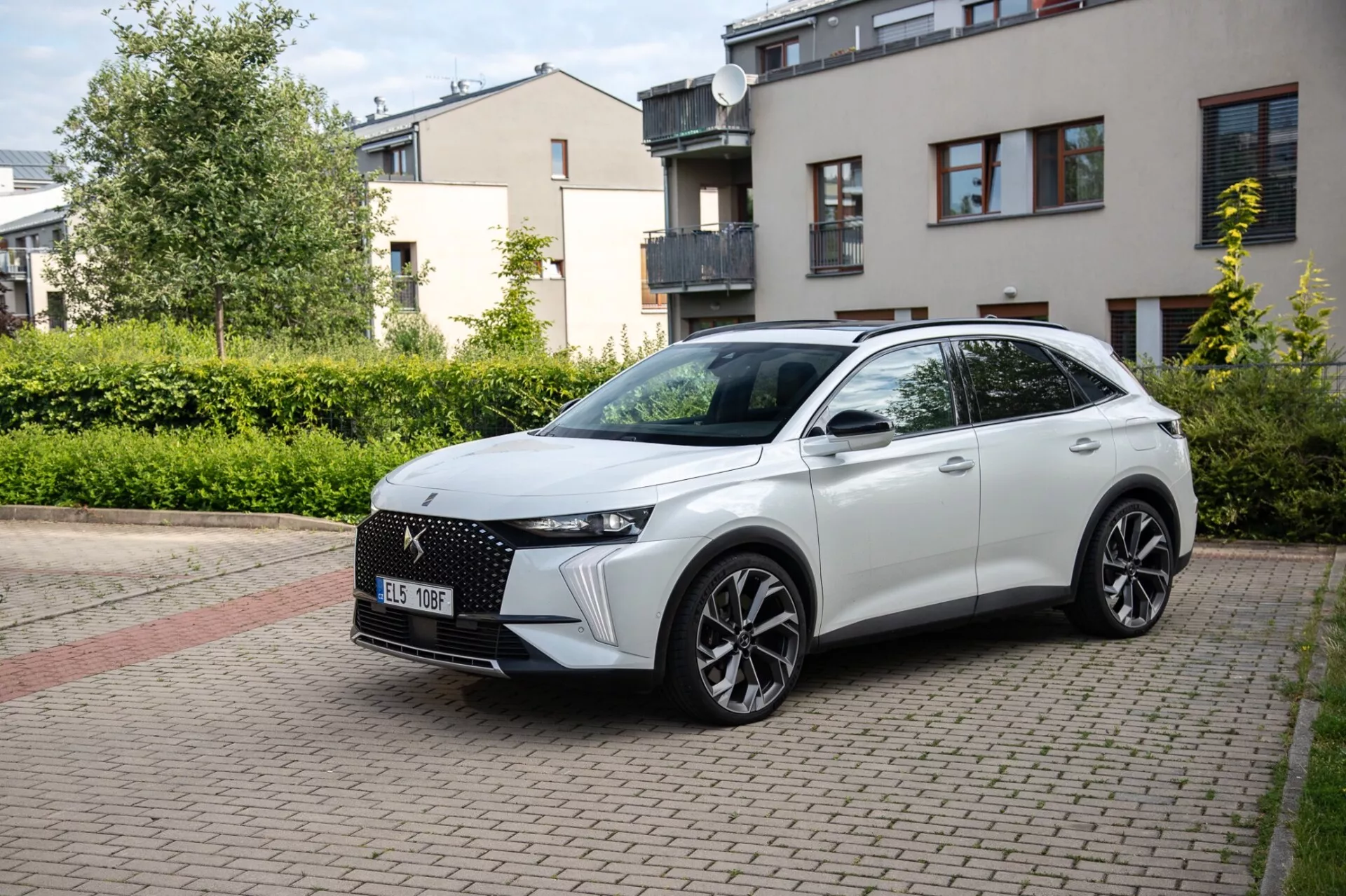 Three-quarter front view of a DS Automobiles DS-7 2022 crossover parked on a brick-paved residential street, showcasing its modern design including a distinctive front grille, sleek LED headlights, sculpted bonnet, large alloy wheels, and a sporty side profile with integrated turn signals on door-mounted side mirrors under natural daylight.