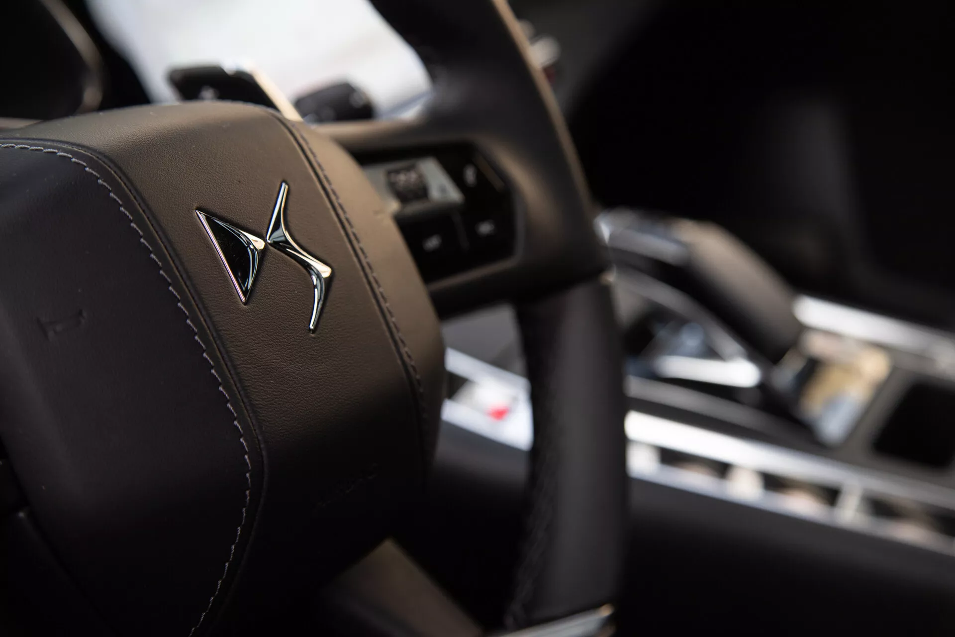 Close-up view of a car's driver seat headrest featuring an embossed logo, with shallow depth of field highlighting the headrest in sharp focus against a blurred interior background showcasing a steering wheel with control buttons and a dashboard with a central display screen.