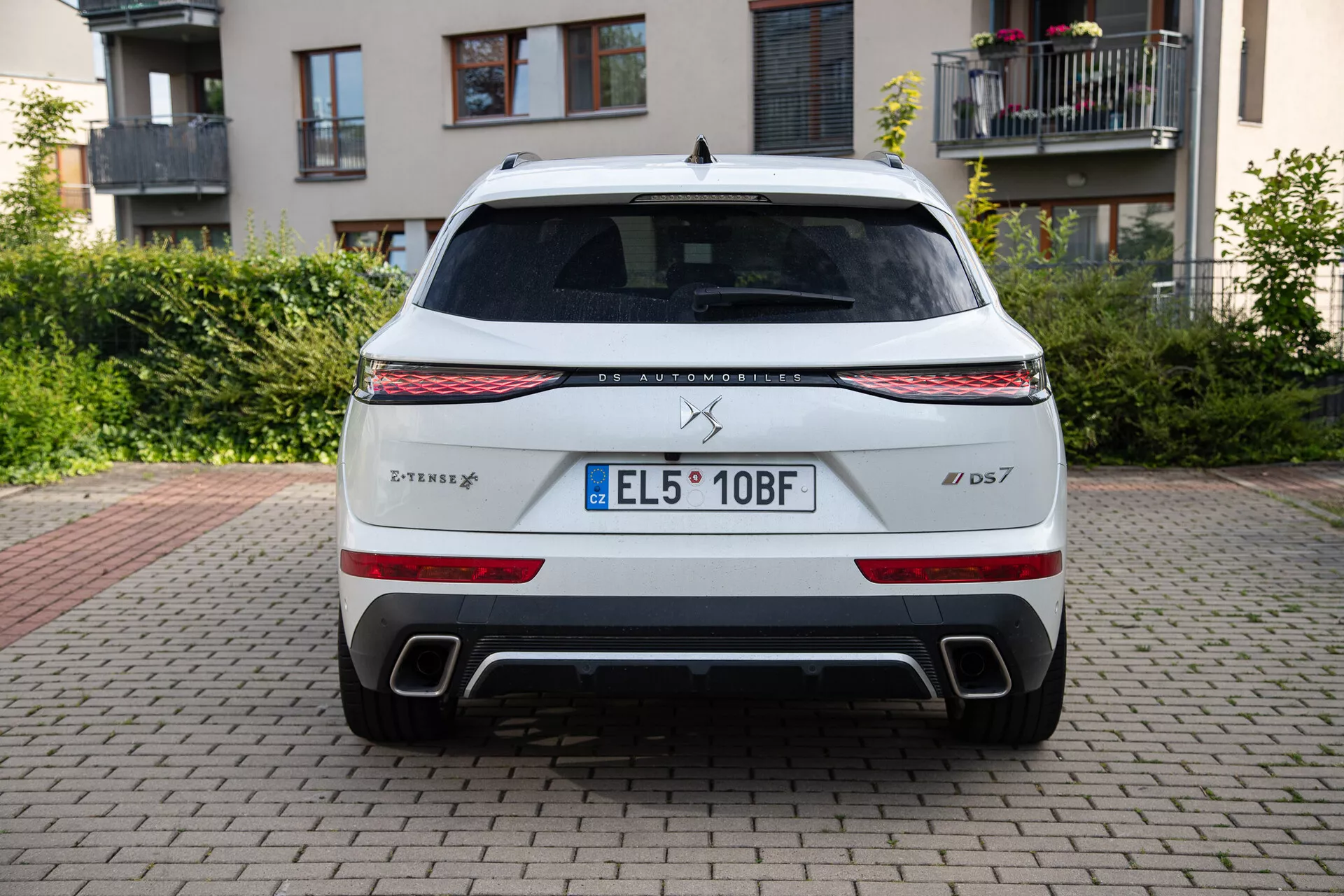 Rear view of a DS Automobiles DS 7 2022 model parked on brick surface with buildings and greenery in background, showcasing LED light strip, taillights, dual exhaust outlets, and EU license plate