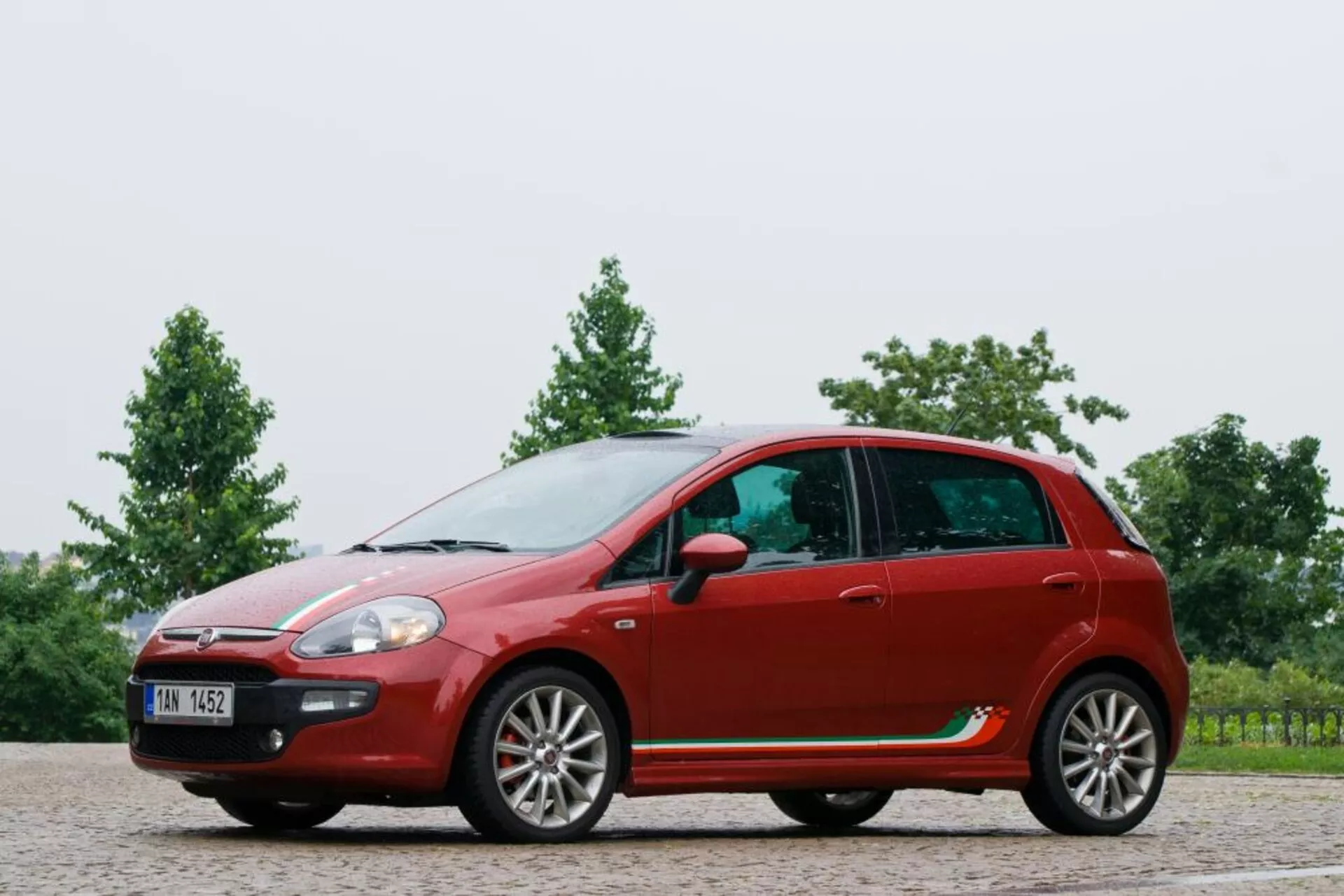 Full side profile view of a 2011 Fiat Punto hatchback in red, showcasing its curved roofline, five-door configuration with alloy wheels, side mirrors on doors, distinct character lines, and plastic molding on a gravel surface with greenery background.