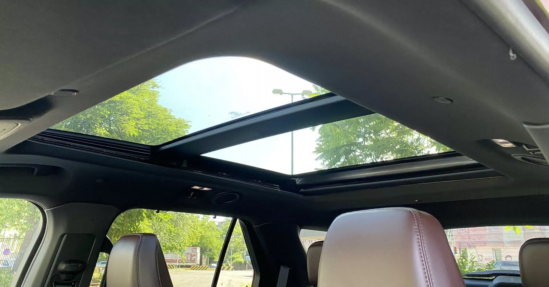 Interior view of a 2019 Ford Explorer focusing on the panoramic sunroof from the rear seating area. The sunroof extends from the front towards the rear with a large glass panel that allows ample natural light, complemented by an open retractable sunshade. The front part of the roof includes overhead console controls and a reading light. The dark headliner contrasts with the lighter leather seats, showcasing a contemporary design and luxury interior features.