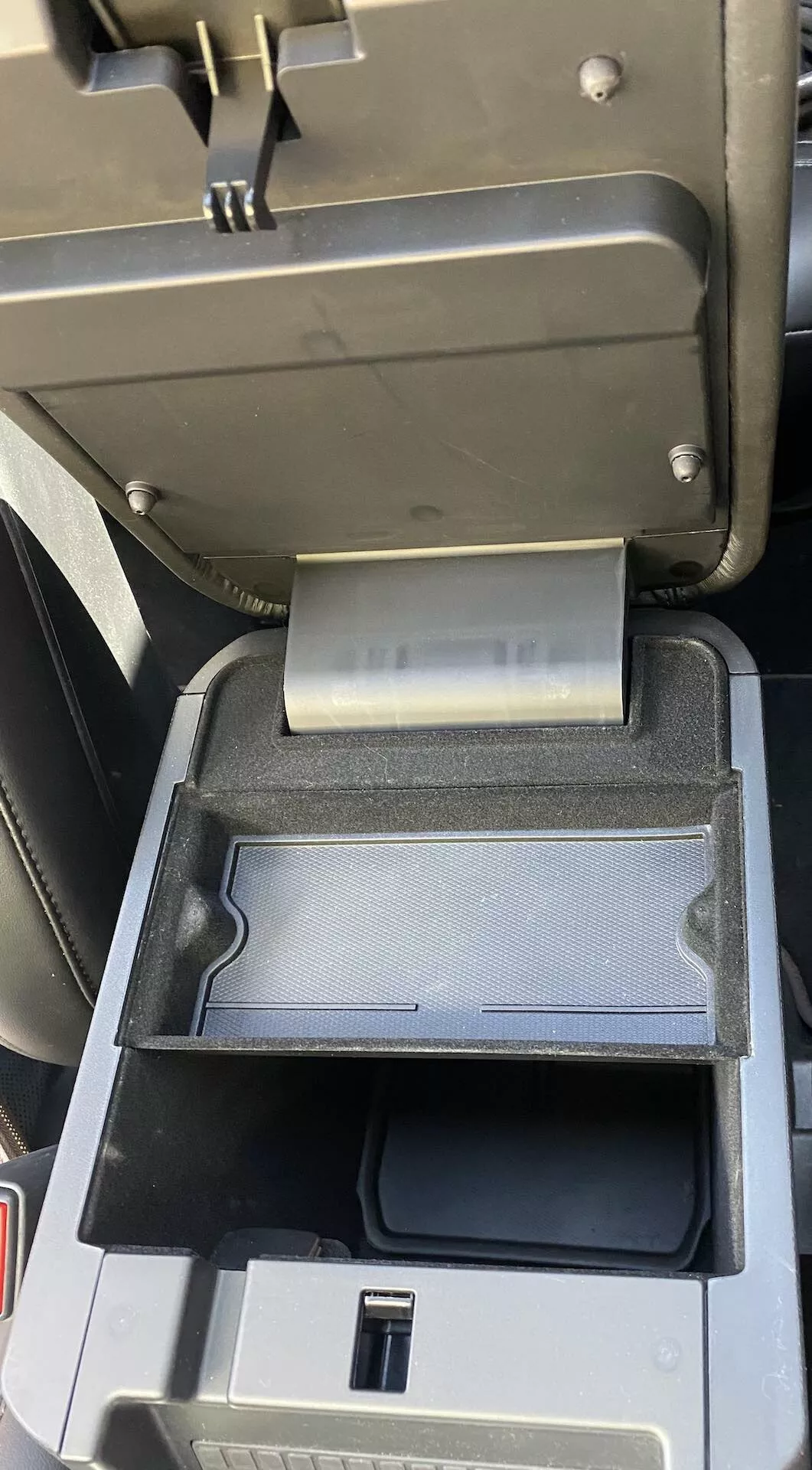 Interior close-up of a 2019 Ford Explorer center console detail showing open center armrest with sliding secondary tray pushed to the back, revealing USB port and deeper storage compartment in black plastic finish.