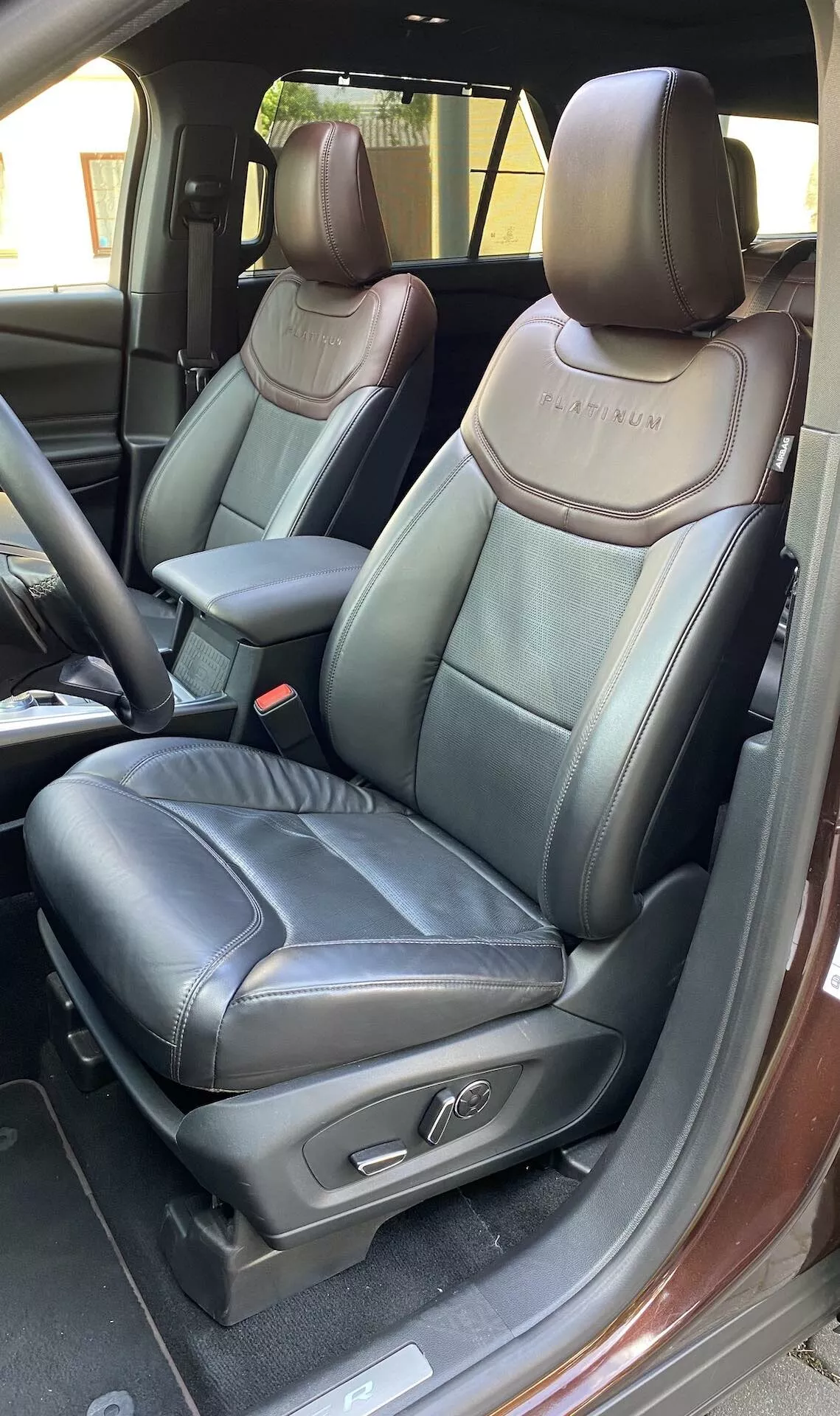 A detailed view of the interior of a 2019 Ford Explorer, focusing on the driver's seat with the door open. The photograph highlights the black leather upholstery with contrasting stitching, an embossed logo on the seatback, adjustable headrest, seat control buttons, central console armrest, visible seat belt release button, internal door lock mechanism, and metallic door sill trim.