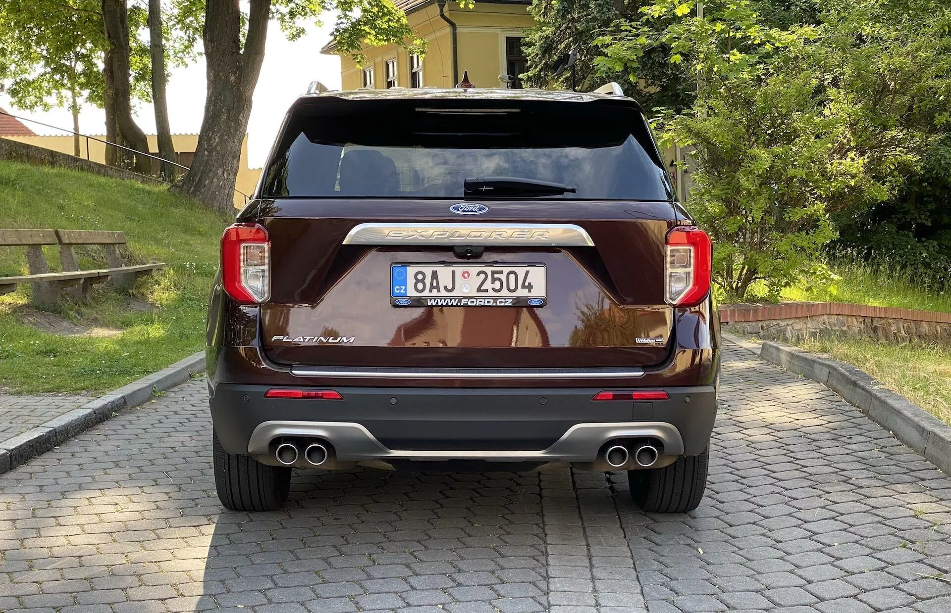 Rear view of a 2019 Ford Explorer SUV. The vehicle showcases a sleek rear design with tail lights on either side, a Ford emblem centered below the rear window, a high-mounted stop lamp, and dual chrome exhaust tips. Additional features include a centrally located license plate with an overhead chrome strip, rear sensors in the bumper suggesting parking assist, and a possible hands-free tailgate system, all captured from a slightly low angle.