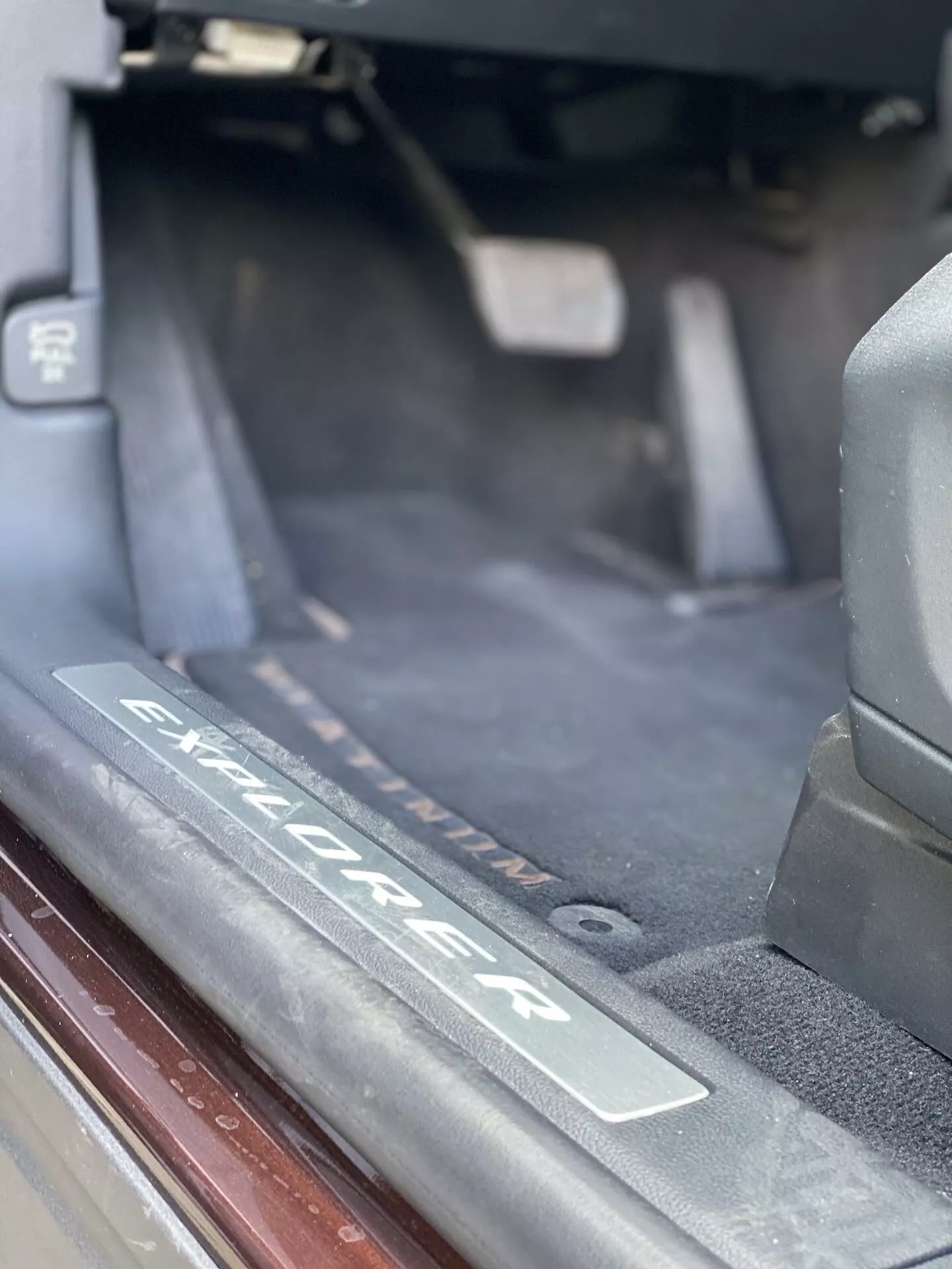 Close-up interior detail of a 2019 Ford Explorer SUV, specifically showing the door sill area with a brushed metal plate etched with the 'Explorer' logo. The image highlights the threshold where passengers step in, featuring dark floor carpeting, weather stripping, and soft natural lighting.