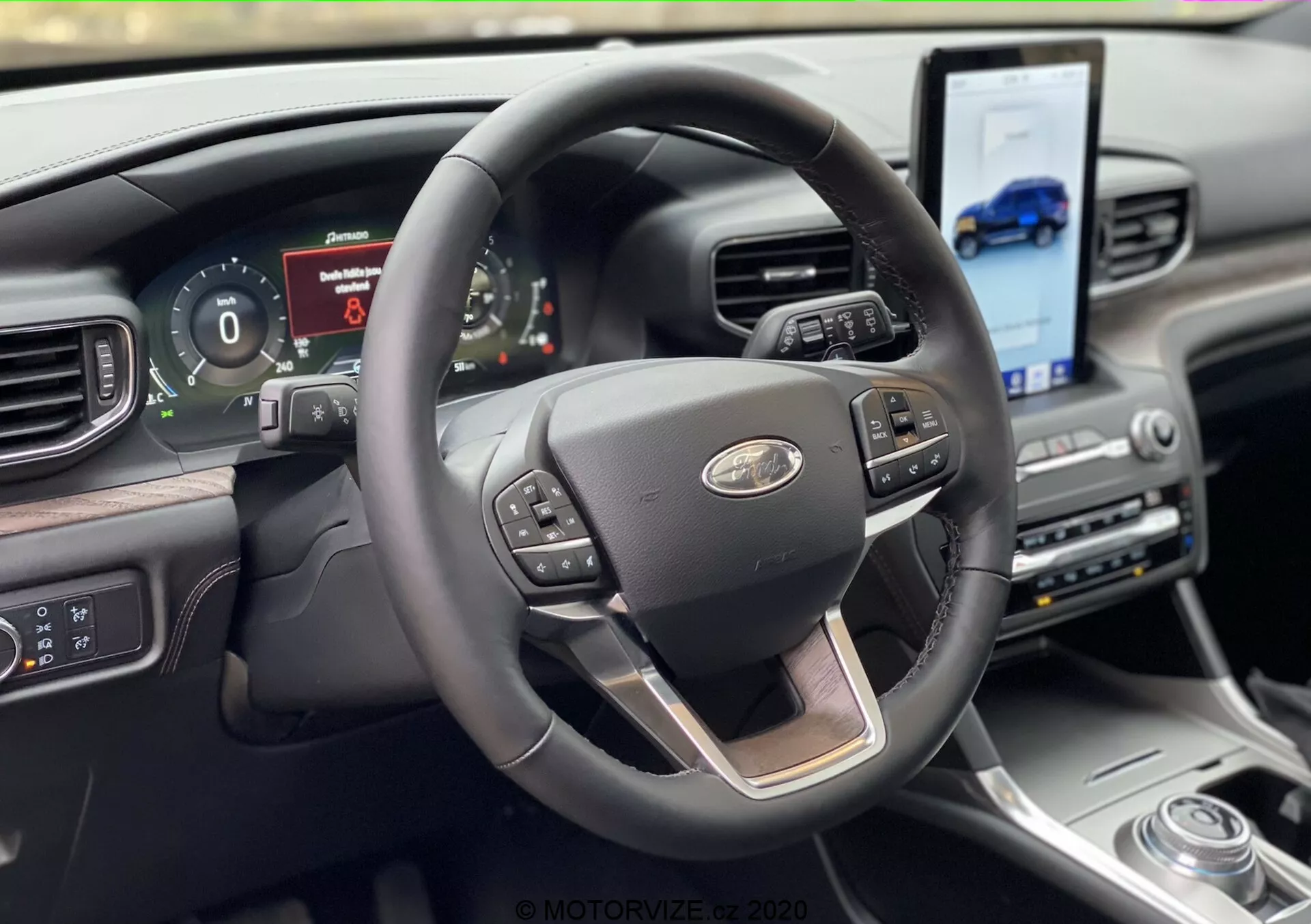 Interior view of the 2019 Ford Explorer from the driver's seat perspective, featuring a modern three-spoke steering wheel with chrome accent and multifunction controls for cruise control, media, and voice commands. Behind the wheel is a digital instrument cluster displaying vehicle information. The dashboard includes a large touchscreen angled towards the driver for navigation and media access, complemented by a rotary gear selector and upscale materials.