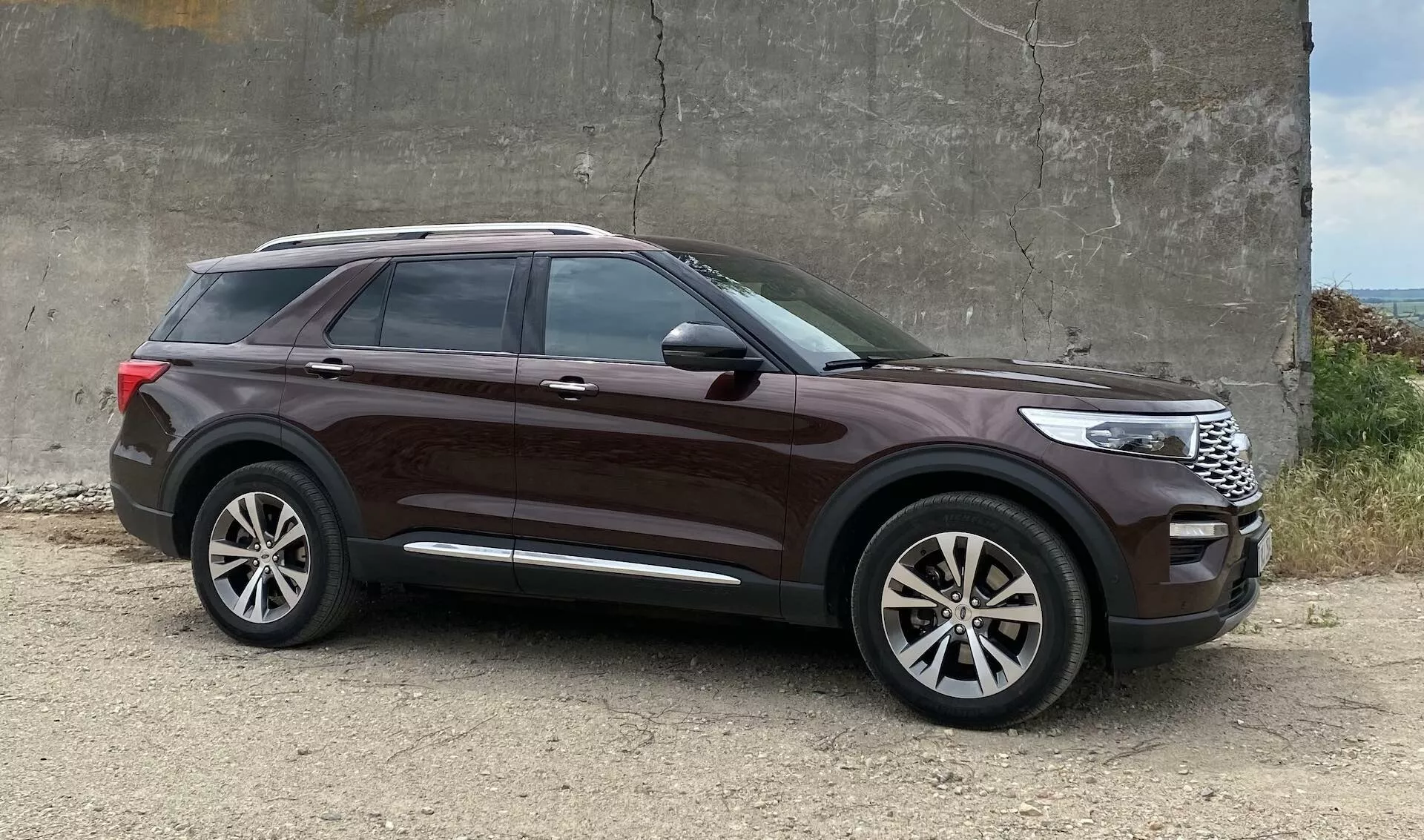 2019 Ford Explorer SUV parked on rough gravel near a concrete structure, side profile angle highlighting design elements like large alloy wheels, high beltline, tinted windows, body-colored door handles, chrome window strip, sleek body lines, integrated turn signal door mirrors, deep metallic paint, and noticeable ground clearance.