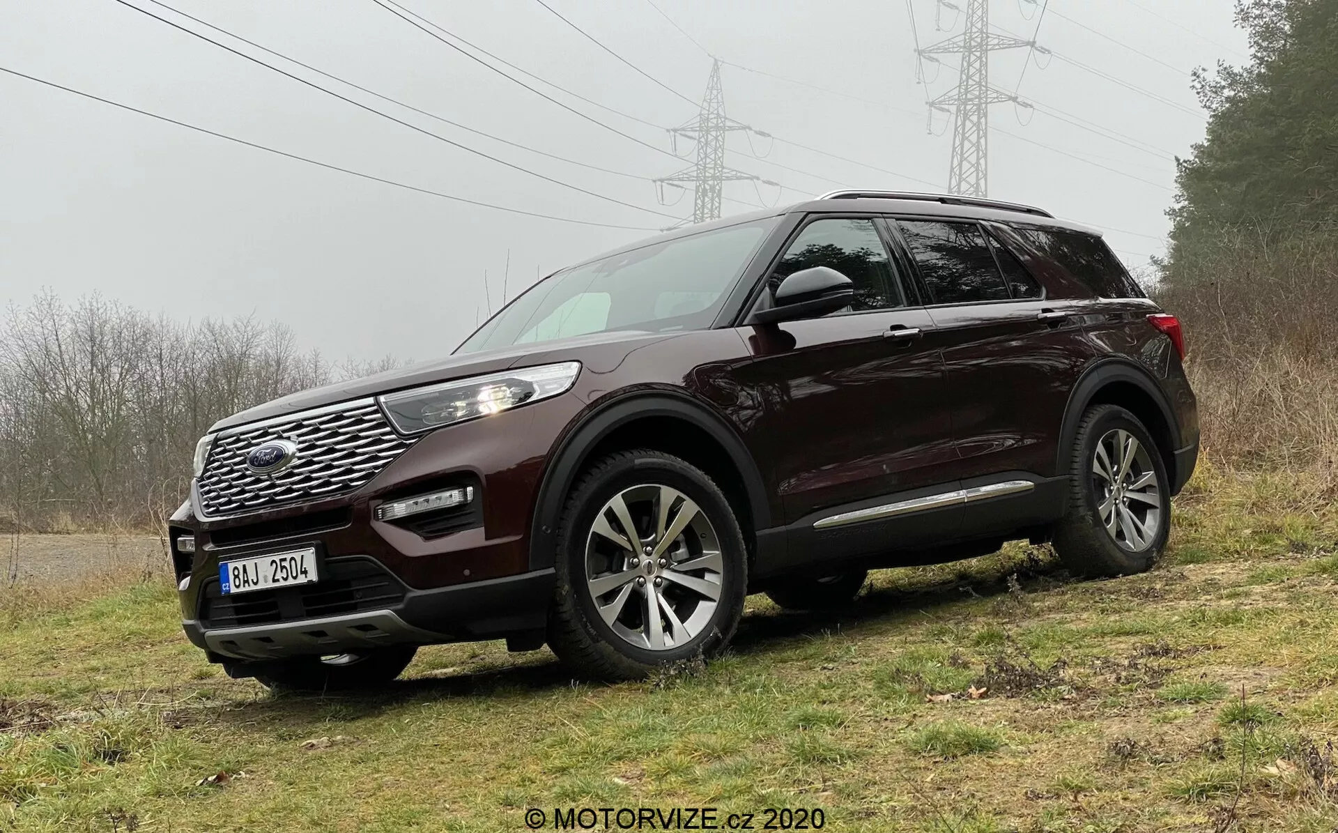 Image of a dark-colored 2019 Ford Explorer SUV parked on uneven grassy terrain, highlighting its rugged and off-road capability. Captured from the front three-quarter angle, the SUV's design elements such as a prominent grille with a geometric pattern, brand emblem, wide air intake, fog lights, sleek wrap-around LED headlights with metallic accents, and multi-spoke alloy wheels are vividly visible. Features also include body-colored mirrors with integrated turn signals and roof rails, suggesting practicality and sophistication.