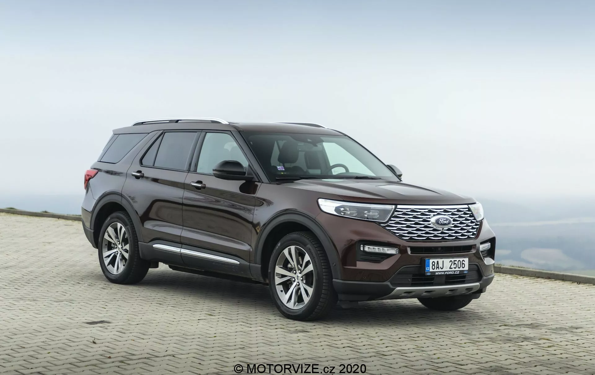 Image of a 2019 Ford Explorer SUV captured from a three-quarter front view angle, highlighting both the side profile and the front fascia. The low camera angle emphasizes the vehicle's robust stance and prominent front grille. The car's full length is visible, showcasing the design lines, wheel design, and contours of the doors and fenders. The wheels are turned slightly to the right, giving a sense of motion. Prominent features include a front grille with horizontal slats, sleek headlights that integrate with the grille, and a front bumper with visible sensors. Side mirrors appear to house integrated turn signals, and the roofline slopes slightly towards the back with visible roof rails. The paint finish is a deep metallic burgundy. The vehicle is parked on a paved surface with a blurred natural background, suggesting a high elevation with a cloudy sky.