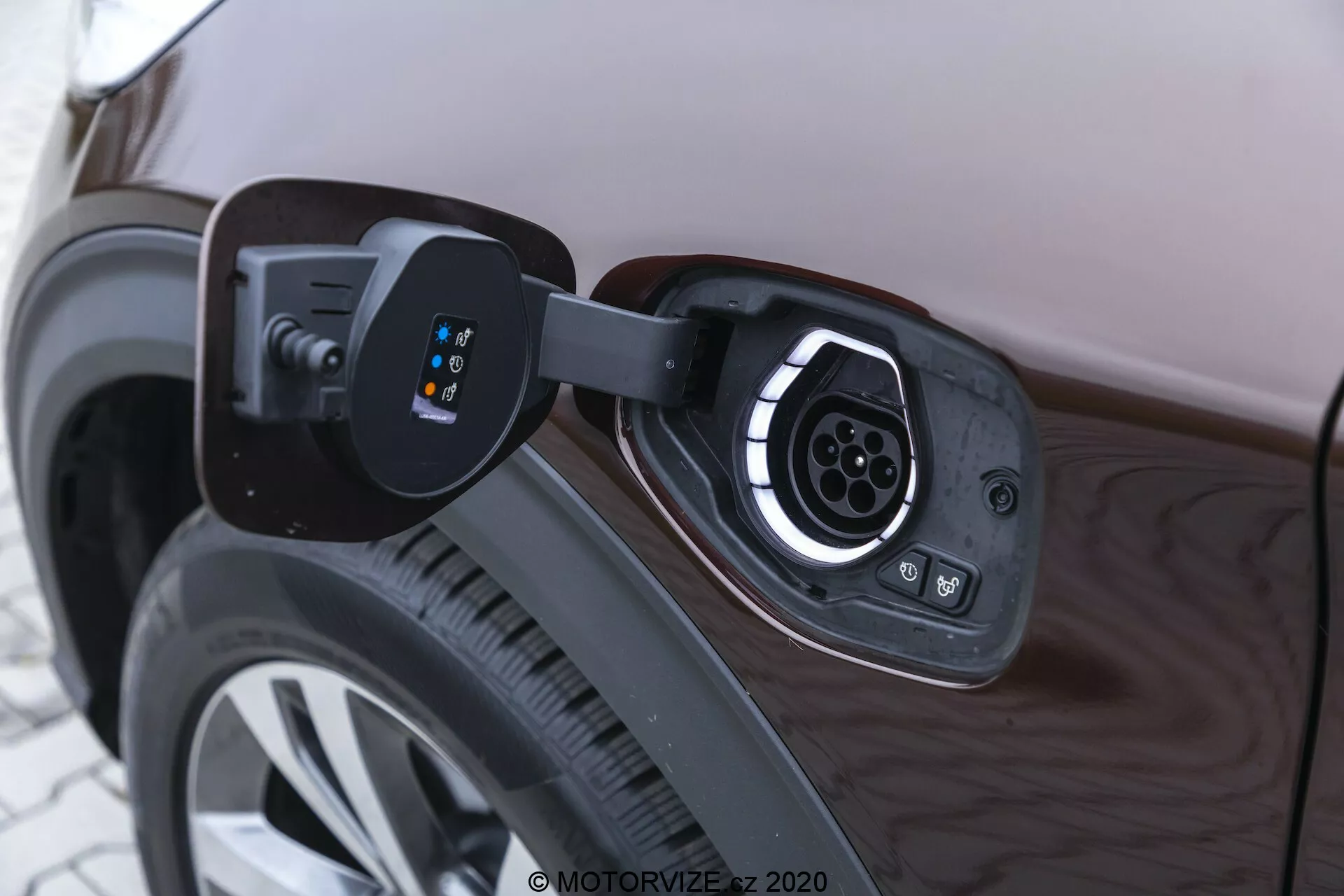 Close-up view of an electric vehicle's charging port on the rear quarter panel near the rear wheel. The open cover reveals the socket with multiple connectors typical of a fast-charging Type 2 or similar standard, featuring several pins in a circular pattern. LED indicators to the left of the socket show charging status. The image focuses on the technology of the charging system, with the car's side panel and wheel rim reflecting ambient light.