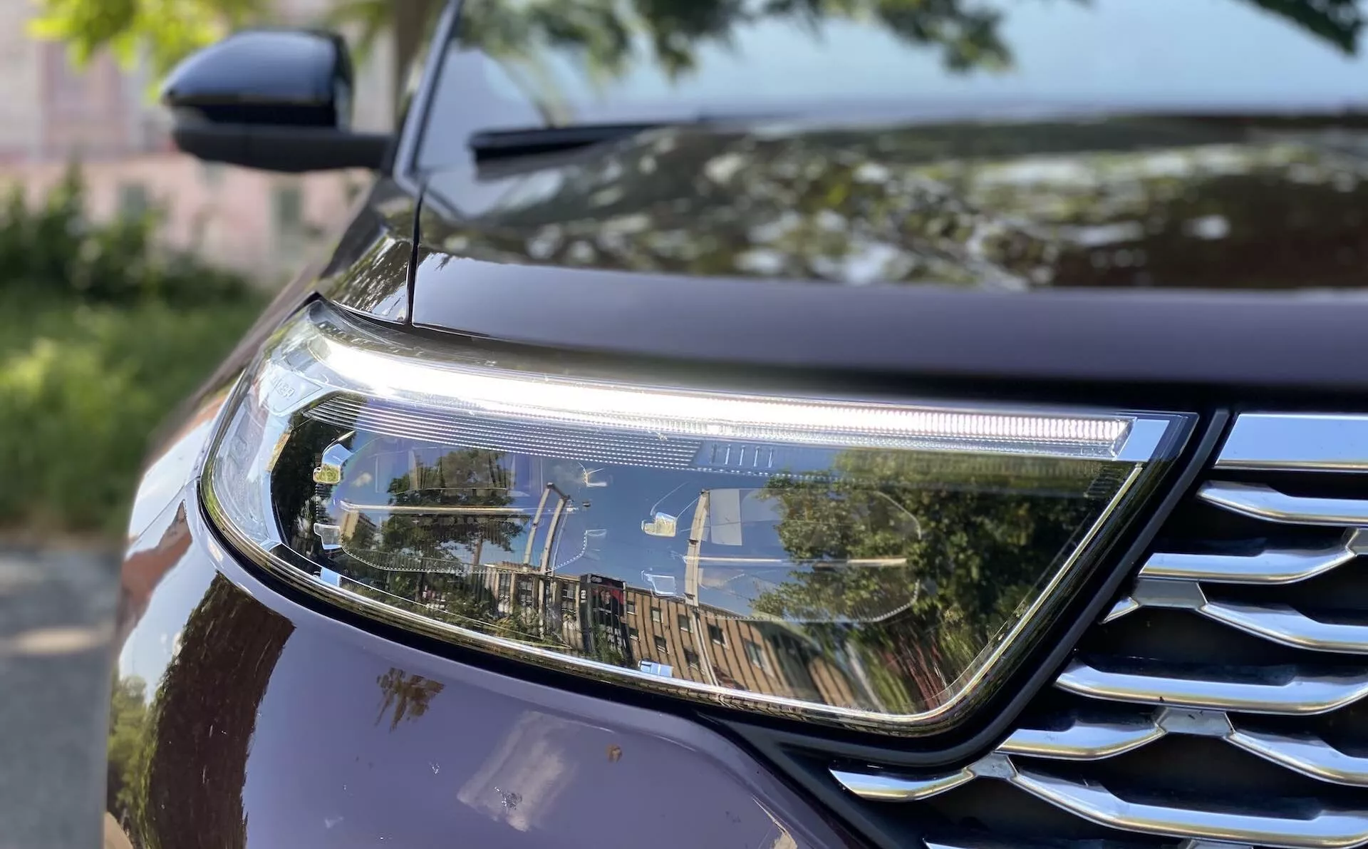 Close-up of the 2019 Ford Explorer's front section showcasing the right headlight and a portion of the grille. The headlight features a modern LED or HID design with a horizontal DRL, while the grille has chrome accents with horizontal slats. The car's dark metallic paint reflects surroundings, highlighting its outdoor setting. The image also captures part of the fender and hood, emphasizing aerodynamic contours.