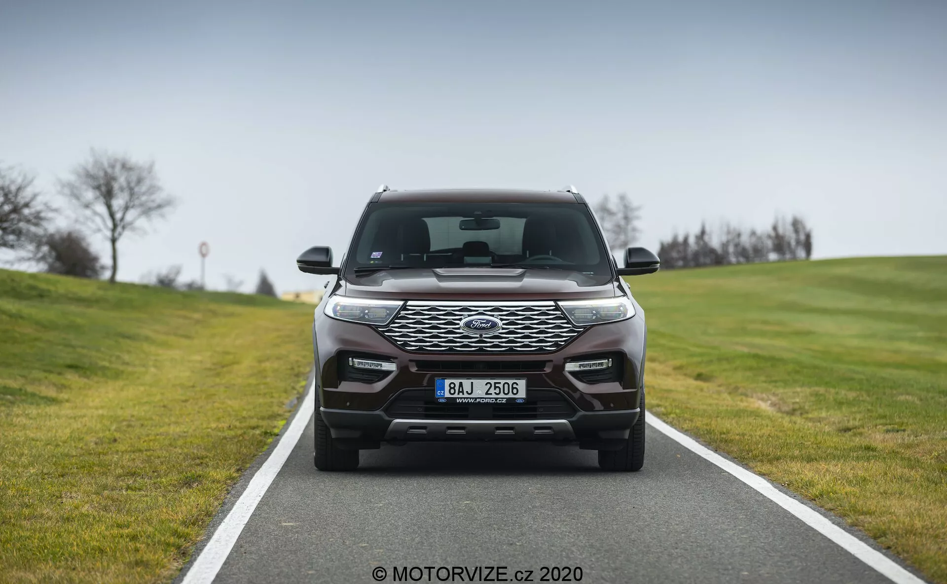 A front-facing image of a modern car, occupying the central frame on a two-lane road, showcasing a sleek headlight design with LED daytime running lights, a prominent honeycomb-pattern front grille, and advanced aerodynamic contours on the bumper, likely housing parking sensors and additional lighting.