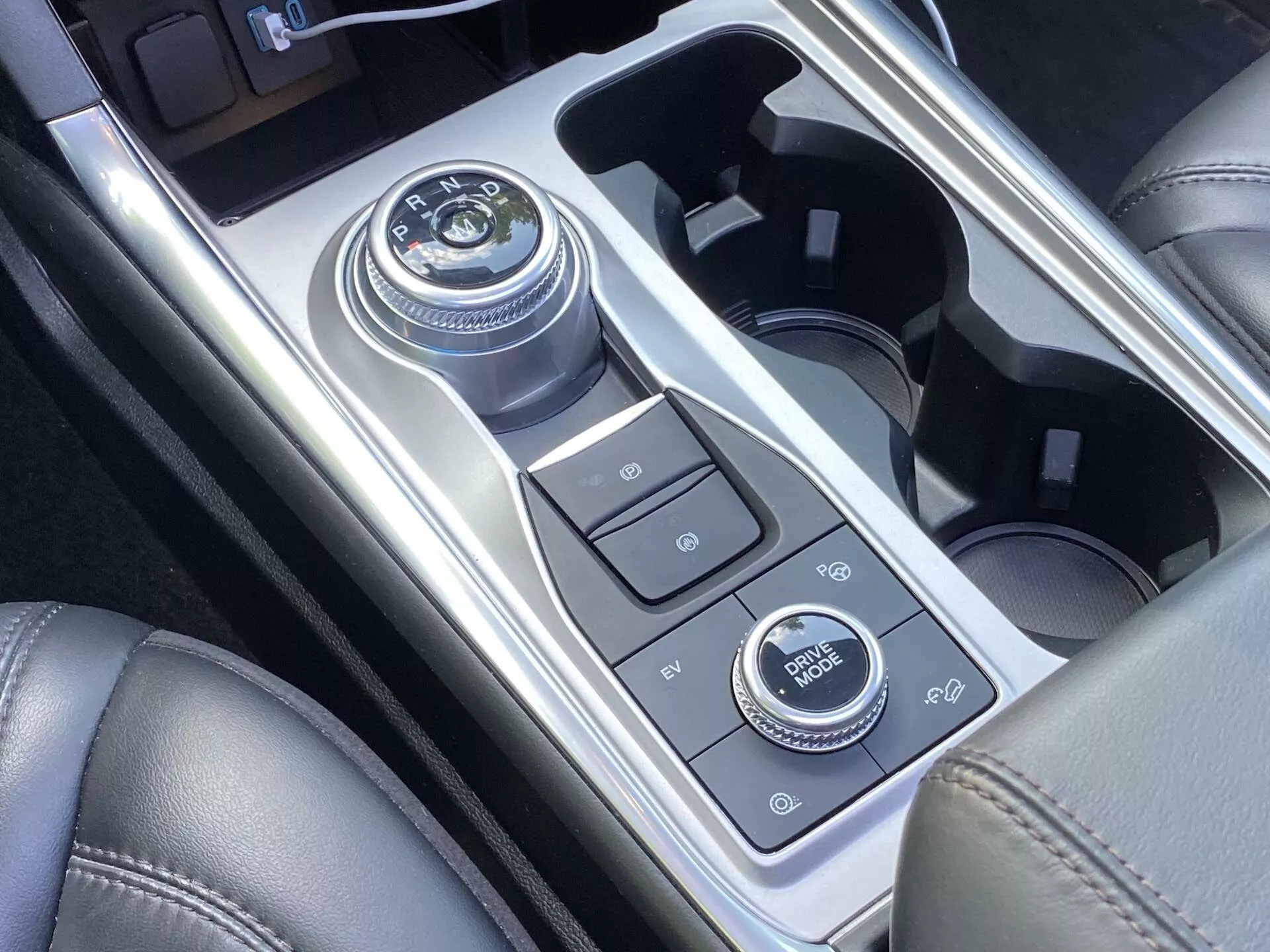 Close-up view of the 2019 Ford Explorer's center console area featuring a rotary dial gear shift, various control buttons, cupholders, and infotainment system dial, all surrounded by high-gloss metallic and matte-finish sections suggesting modern luxury and advanced technology.