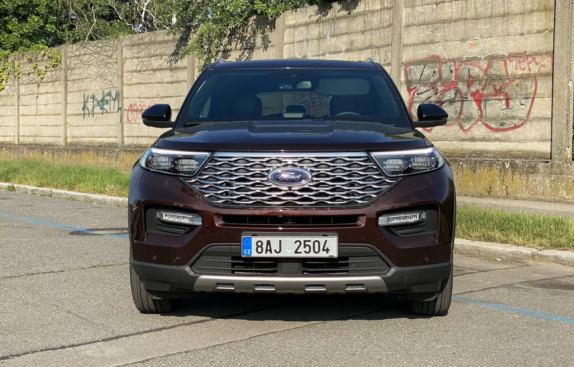 Frontal view of a 2019 Ford Explorer SUV captured from a level angle, showcasing the vehicle's modern design. The prominent front fascia features a large, chrome-finished grille with a hexagonal mesh pattern and centrally located manufacturer's emblem. Sleek, horizontally elongated LED headlights and a color-matched front bumper with lower air intakes and parking sensors highlight the technological advancements. The hood displays a contoured design with raised lines, while the fenders and integrated turn signal side mirrors add a muscular appearance. The SUV, painted in a rich maroon or dark red color, is parked on a paved area with a concrete curb and a graffiti-covered wall in the background, accentuating an urban setting.