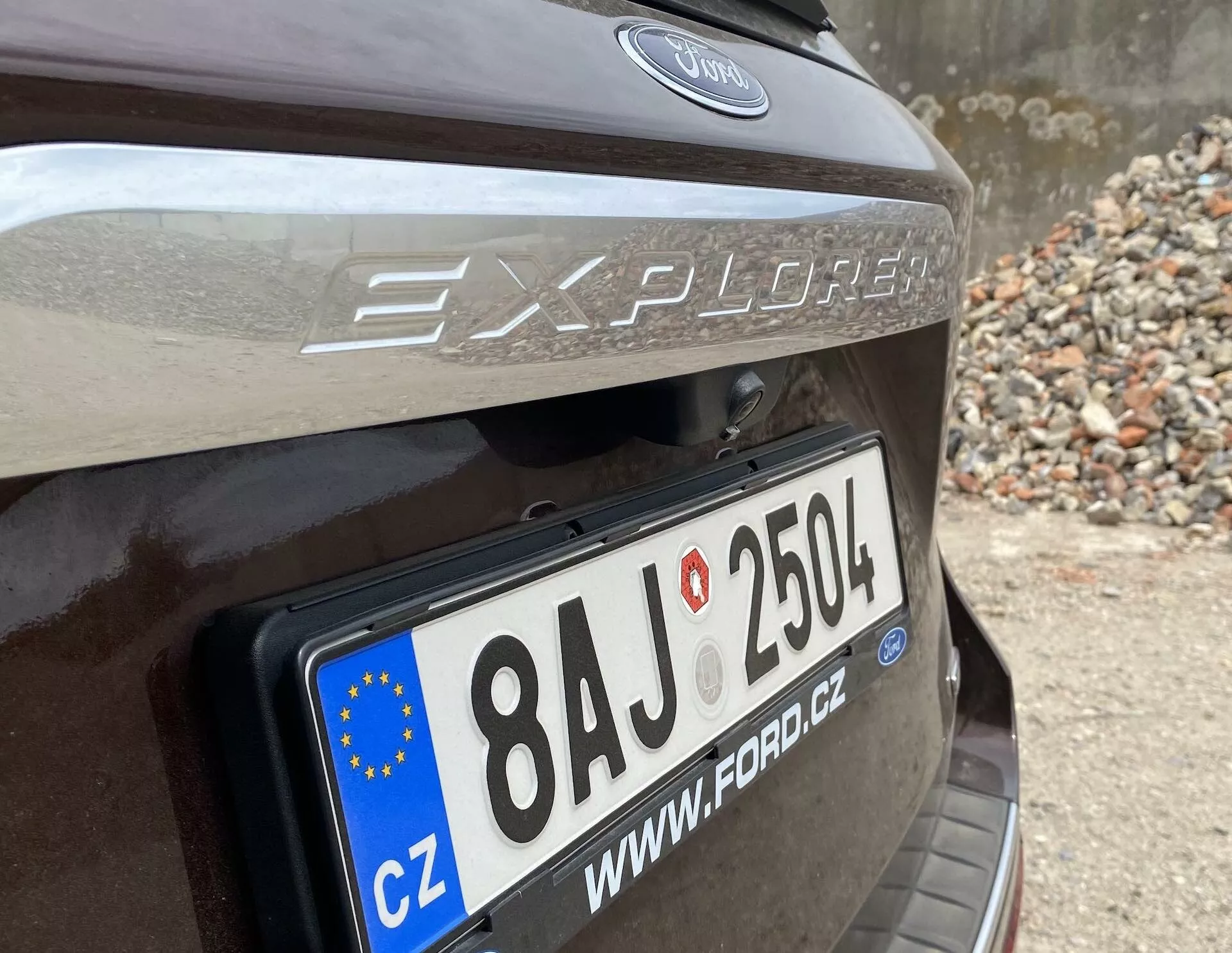 Close-up view of the rear section of a 2019 Ford Explorer, focusing on the liftgate area with a prominently displayed European Union license plate reading '8AJ 2504' and a 'CZ' country code. The 'Explorer' model name and Ford logo are above the plate, with reflective trim visible above the license plate. Car appears to be dark brown or dark grey, parked in an area with stones or rubble.