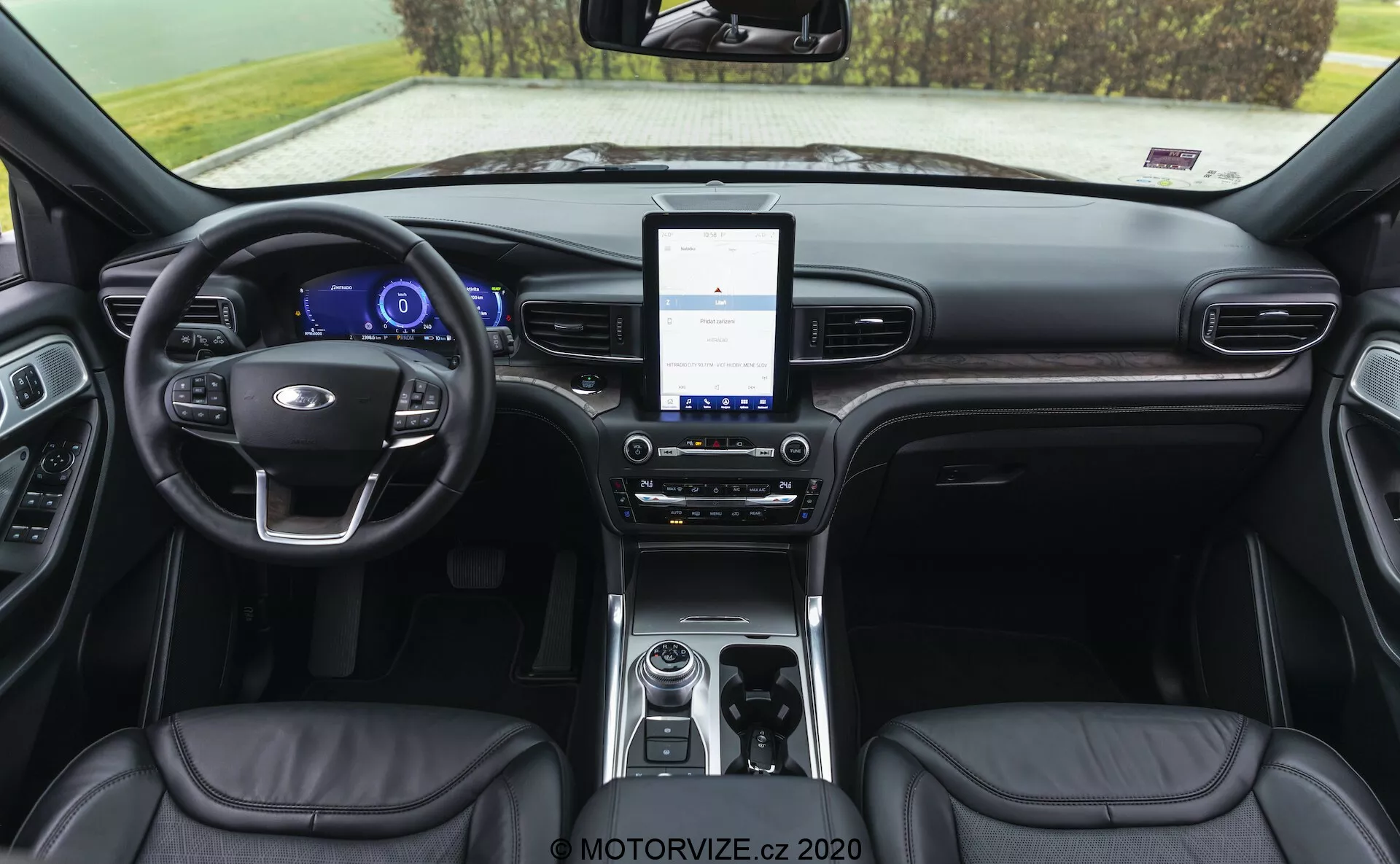 Interior cabin of a 2019 Ford Explorer showcasing the dashboard area from a rear-facing angle. The view includes a leather-wrapped steering wheel with mounted controls, digital instrument cluster, large touchscreen infotainment system, dual-zone climate controls, rotary-style gear selector, storage compartment with connectivity options, leather-upholstered seats with visible stitching, and partially visible door panels with controls.