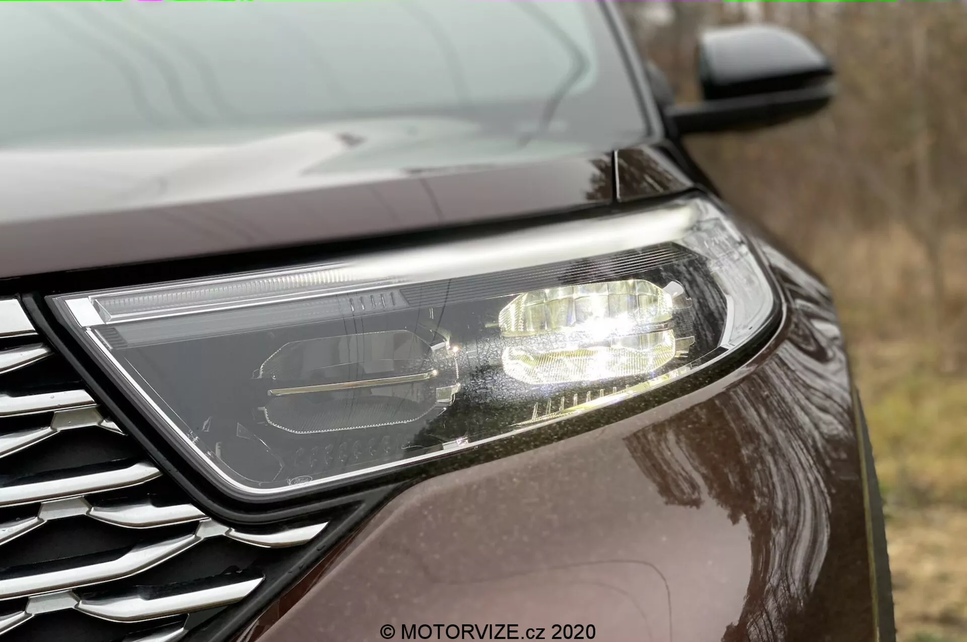 Close-up side view of a Ford Explorer 2019 front end. Features prominent modern headlight suggesting LED or HID technology with active daytime running lights. Partially visible grille with a metallic-finish badge, positioned at a low angle. The car has a dark, reflective paint finish, indicating a metallic or pearlescent quality, in an outdoor setting.
