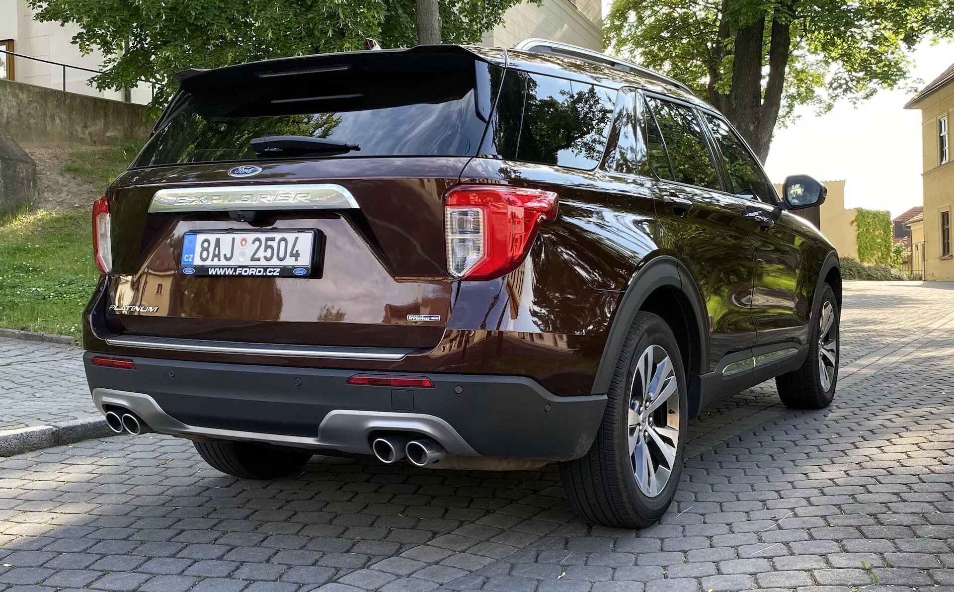 2019 Ford Explorer modern SUV from rear three-quarter perspective showcasing taillight design, rear bumper with parking sensors, dual exhaust pipes, multi-spoke alloy wheels, and dark metallic paint finish, parked on cobblestone surface in urban setting with foliage and buildings in background.