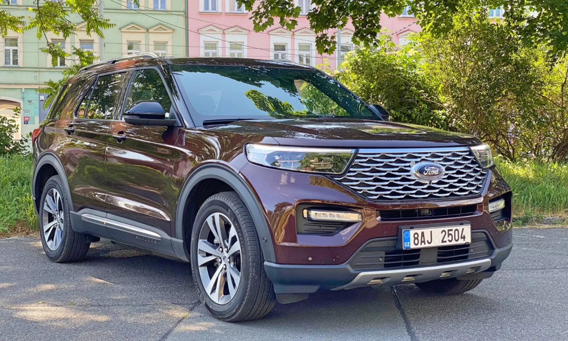 Three-quarter front view of a 2019 brown Ford Explorer SUV, showcasing its side profile and front fascia. The car is parked on an urban roadside with buildings and trees in the background. Key features visible include a prominent front grille with horizontal slats and central emblem, modern LED daytime running lights, fog lights integrated into the lower bumper, large multi-spoke alloy wheels, side mirrors with turn signal indicators, tinted windows, roof rails, contoured body lines, and raised ground clearance.