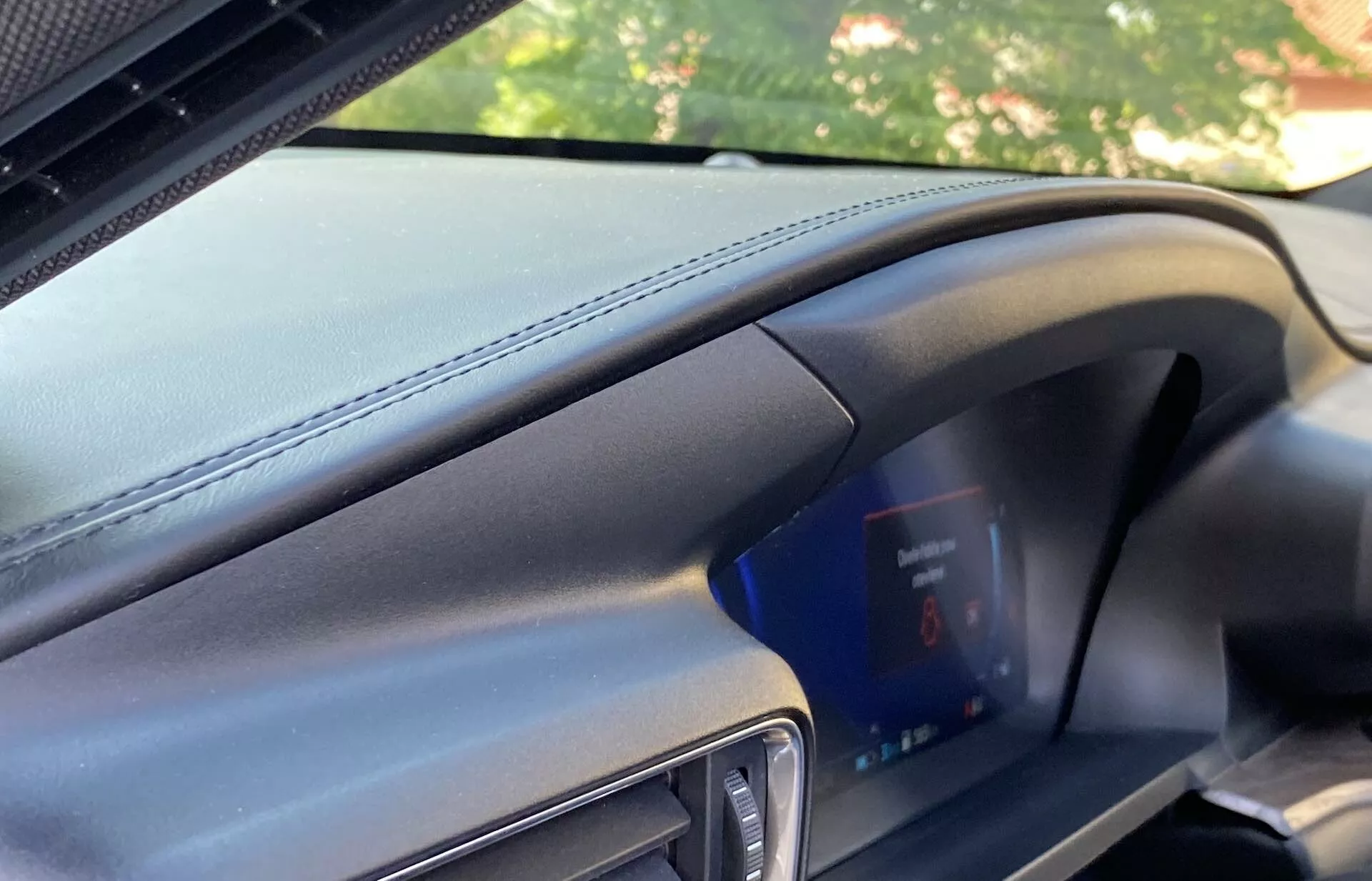 Interior view of a Ford Explorer 2019 from the passenger's perspective showing a modern leather-like textured dashboard with prominent horizontal stitching and a digital instrument cluster featuring a blue screen with red warning indicators. The dark grey and black color scheme and partial view of the car's door frame suggest a premium vehicle taken in a well-lit environment.