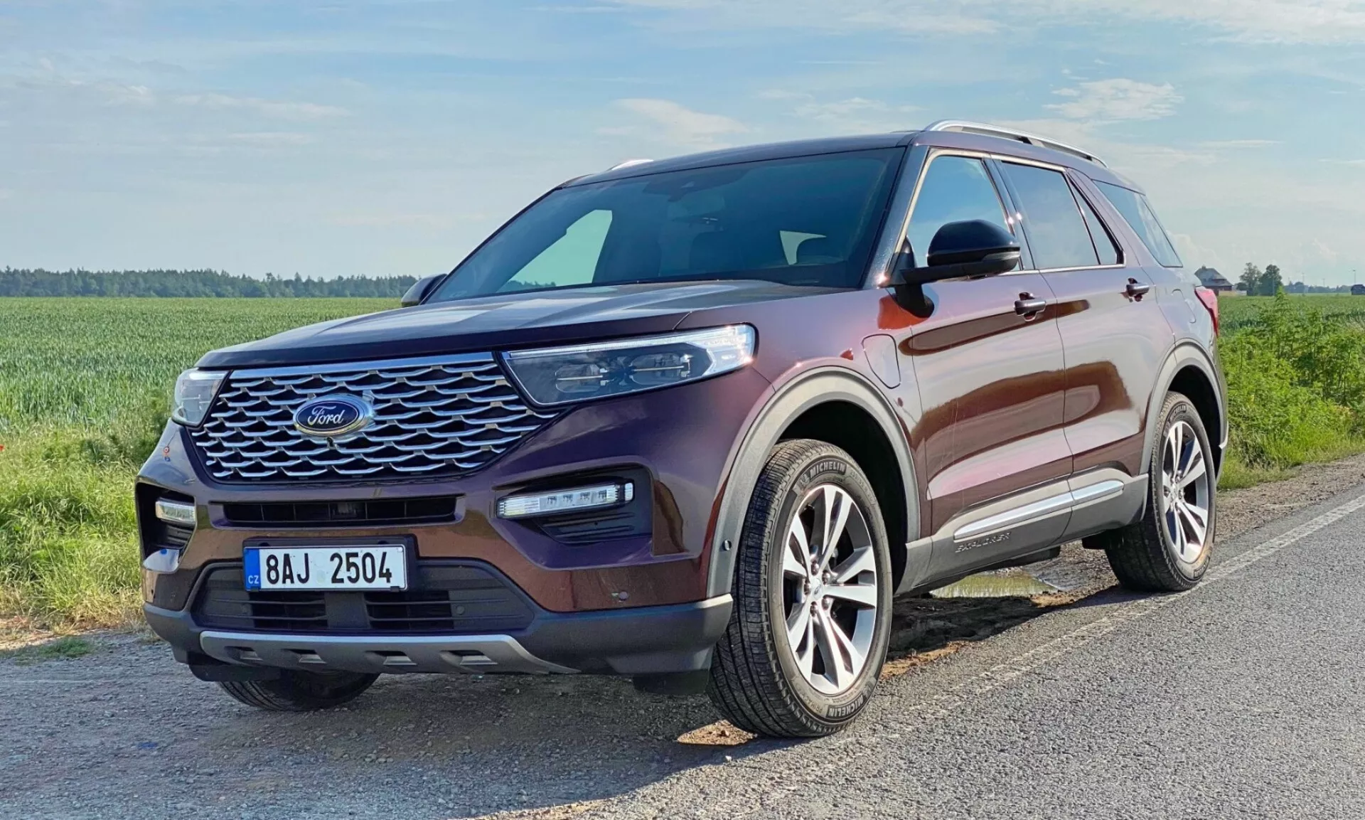 Three-quarter front view of a 2019 Ford Explorer SUV. The image showcases the vehicle's front grille with a mesh-like pattern, sleek elongated headlights with visible LED daytime running lights, a sculpted hood with raised contours, alloy wheels with a multi-spoke design, and color-matched side mirrors with integrated turn signals. The windows display a chrome or silver-colored trim. The vehicle is metallic maroon and positioned on an asphalt road in a grassy, potentially rural area.