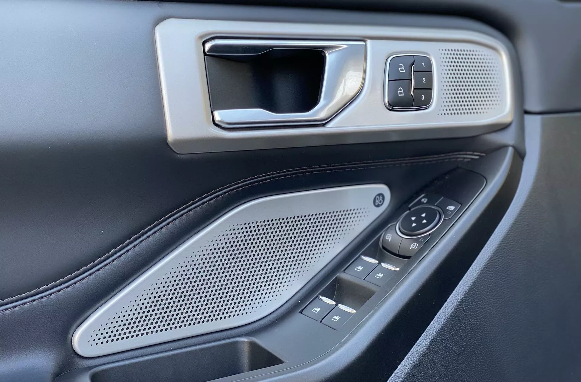 The image captures the driver's side interior door panel of a 2019 Ford Explorer. The focal point from an elevated, slightly angled perspective showcases the metallic-looking door handle embedded in a bezel, alongside the lock/unlock button set within a fine-grained textured panel. Below, an oval-shaped speaker grille with round holes, several control buttons and switches for side mirrors and windows are visible, suggesting power windows for multiple doors. The dark color palette with contrasting metallic elements and visible stitching on a likely leather armrest enhances the luxury feel. Shallow depth of focus highlights the control elements, emphasizing design and placement.