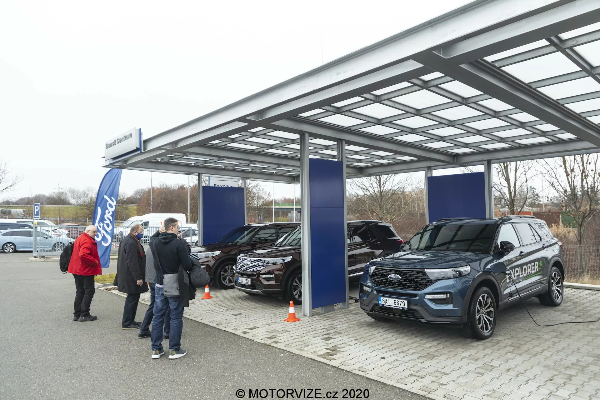The image captures an outdoor car dealership or exhibition event under a metallic, semi-transparent roof with soft, overcast daylight. Three SUVs, including a modern Ford Explorer 2019 in the foreground, are on display with features like front grille, headlights, and side mirrors visible. People and banners suggest an official promotional event, and the Ford logo is discernible in the background.
