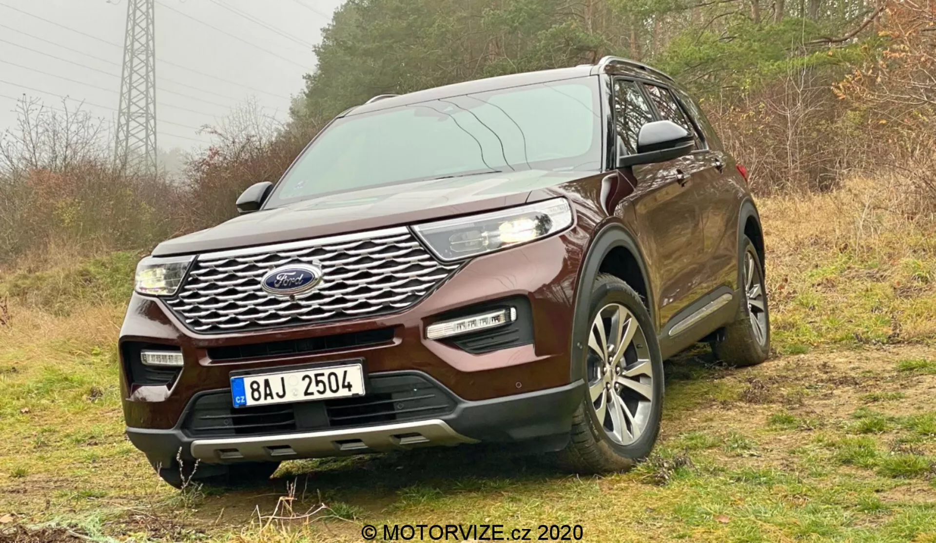 Frontal three-quarter view of a 2019 Ford Explorer SUV taken from a low angle, showcasing its prominent chrome grille, sleek LED headlights, and multi-spoke alloy wheels. The SUV is positioned on an unpaved, vegetative surface hinting at its off-road capability. The front bumper includes fog lights and air intakes, suggesting enhanced functionality. Elements like sensors or cameras possibly indicate advanced safety features. The license plate is censored, and a watermark reads '© MOTORVIZE.cz 2020'. The angle emphasizes its robust stance and technological sophistication.