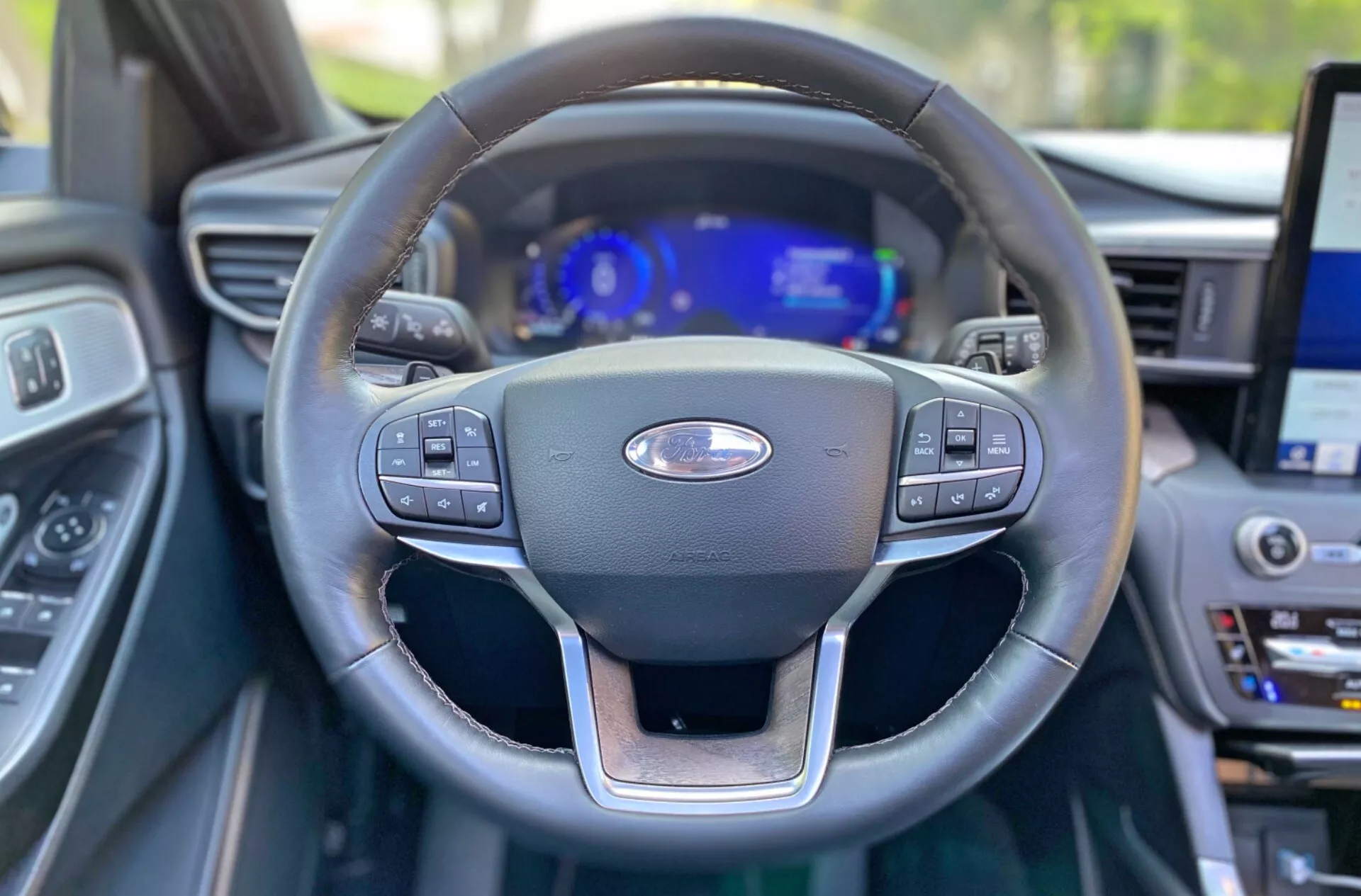 The image shows an interior view of a car focusing on the steering wheel and the dashboard, taken from the driver's seat perspective, slightly angled upward towards the steering wheel. The foreground showcases the steering wheel with control buttons on both the left and right spokes for multimedia system, cruise control, and voice commands. A digital dashboard behind the steering wheel displays the speedometer, tachometer, and other driver information with blue backlighting, indicating an advanced and customizable instrument cluster. Partially out of focus to the far right is an infotainment screen, hinting at the car's central display for settings, GPS navigation, and media playback. The interior design and materials suggest a modern, high-end vehicle aimed at enhanced driver interaction.