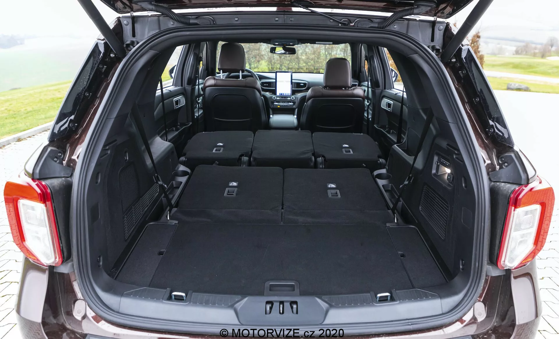 The image shows the open trunk of a 2019 Ford Explorer from a slightly low rear angle, showcasing its spacious cargo area with rear seats folded down. The flat load floor, dark carpeted interior, split 60/40 rear seats, side interior lighting, and visible 12-volt power outlet highlight the vehicle's cargo space and technological convenience features. The photo emphasizes a functional, wide, and deep trunk, with a retracted privacy cover shelf and a blurred exterior indicating a shallow depth of field.