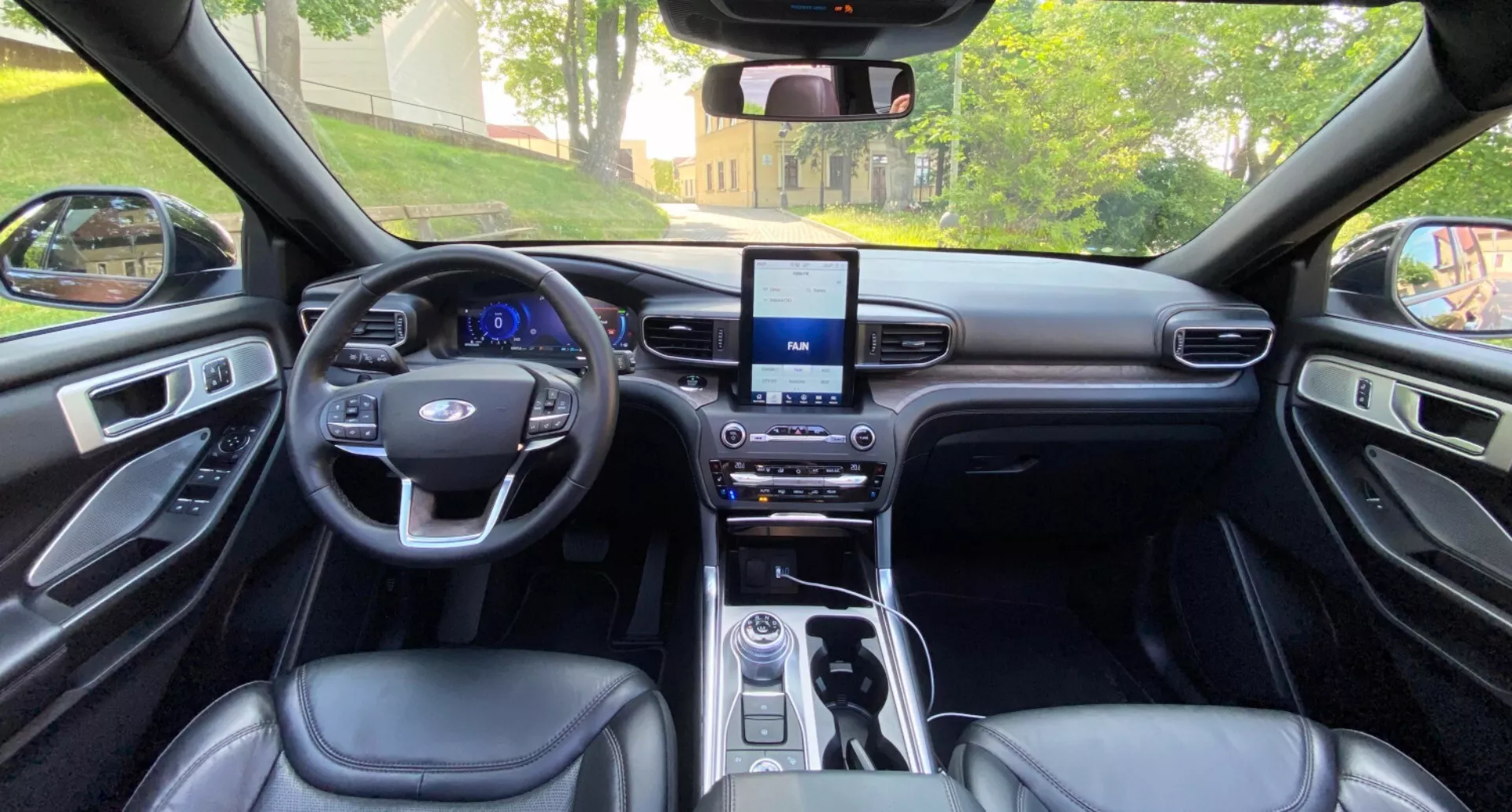 Interior view of the Ford Explorer 2019 showing the perspective from the rear seats looking forward towards the dashboard. The image captures the symmetrical layout including the steering wheel with multifunction buttons, the infotainment system touchscreen, the gearshift console with controls, and the leather-like upholstery on the seats and door panels. The premium interior is highlighted with sleek metallic trims and well-placed illumination.