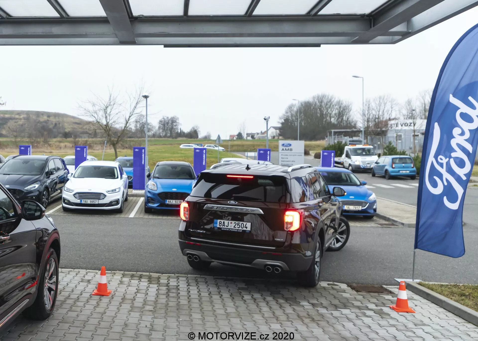A black 2019 Ford Explorer SUV, positioned centrally, is in the process of either backing into or pulling away from an EV charging spot at a charging station. Rear lights are on, possibly indicating reverse or braking, with a visible license plate, dual-sport exhaust system, rear window wiper, and high-mounted rear brake light. The surrounding area shows several electric cars at blue charging poles and orange traffic cones near the SUV. A flag with the Ford logo suggests brand presence. The scene is captured on an overcast day with diffused natural light, highlighting automotive technology and sustainable transport infrastructure.