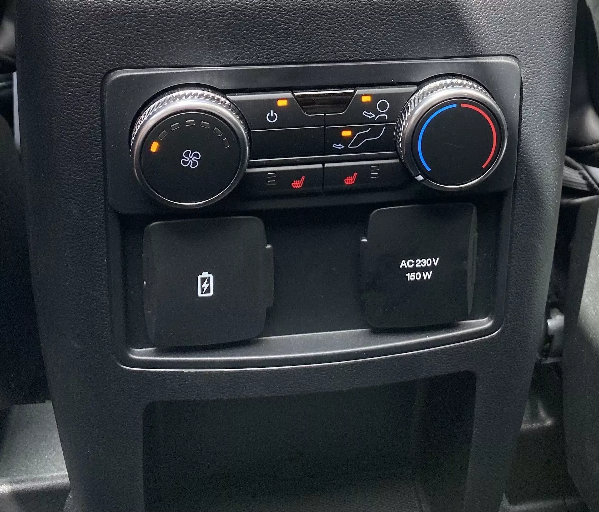 Close-up view of a section of the 2019 Ford Explorer car interior center console, showcasing HVAC and comfort controls. On the left, a round black dial with a fan icon adjusts the blower; next to it, buttons for airflow direction, air recirculation, and windshield defrost functions. A blue and red dial on the right controls temperature. Below, two anti-slip storage compartments, one designed for a key card, and a passenger-side AC power outlet offering 230V and 150W for electronic devices.