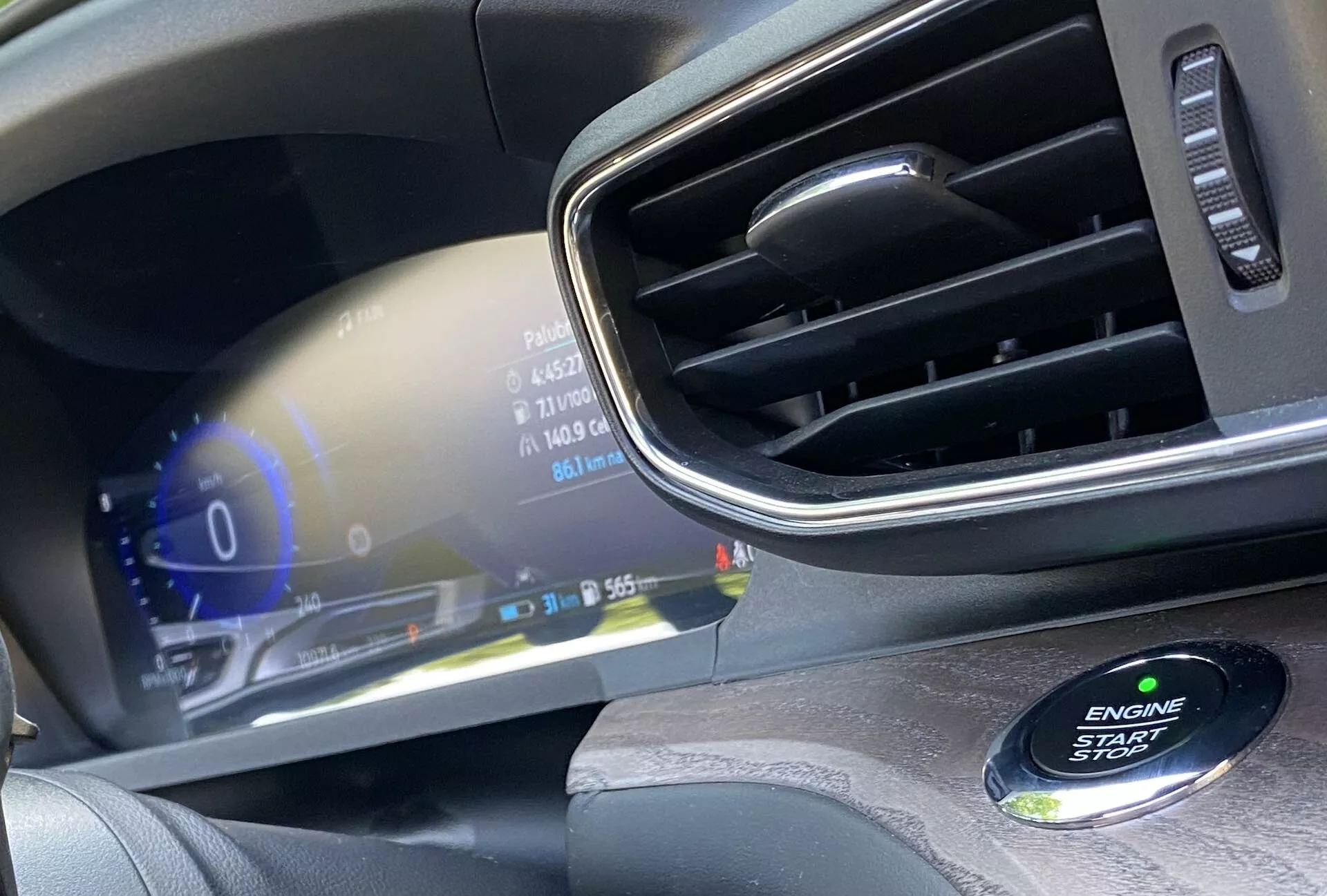 Close-up interior view of a modern car's driver's side dashboard. The image emphasizes the digital instrument cluster displaying vehicle statuses such as speedometer at 0 km/h, fuel gauge, and 31 degrees Celsius outdoor temperature. The sleek, high-resolution display boasts vivid colors and luxurious design. On the right, a chrome-accented air vent is visible, and in the bottom right corner, a slightly out-of-focus ENGINE START STOP button indicates a keyless start system.