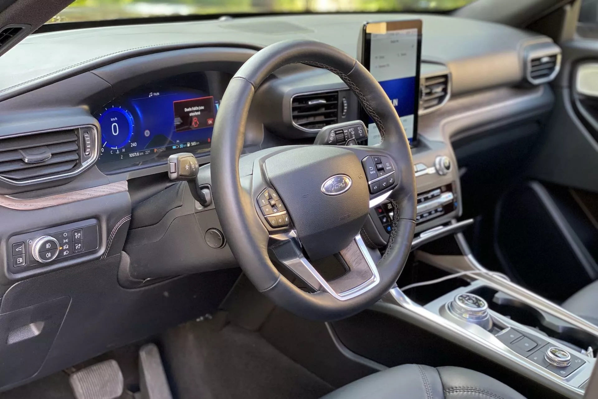 Interior shot of a 2019 Ford Explorer focusing on the driver's area from the front passenger perspective. The steering wheel, featuring multifunction controls for audio, cruise, and hands-free communication, is centered. Behind it, a digital instrument cluster displays customizable gauges and real-time vehicle data. To the right, a vertically oriented touchscreen shows an infotainment system capable of satellite navigation and smartphone integration. Below, multi-zone climate controls and a rotary dial gear shift indicate a contemporary design. The cabin's clean and modern dashboard, though made of premium materials, exudes a minimalist aesthetic.
